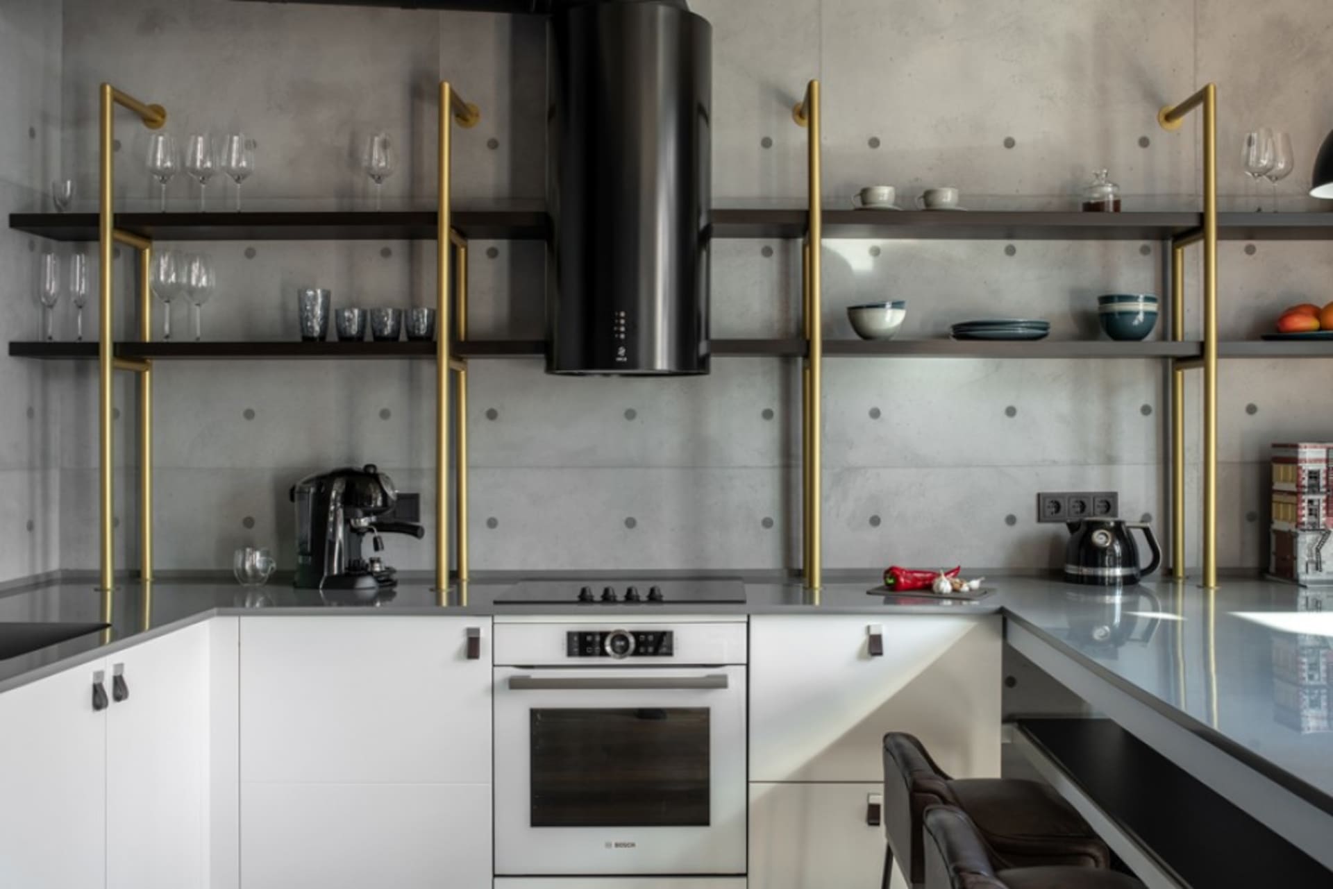 A modern minimalist kitchen with concrete walls, featuring a black and gold shelving unit, white cabinetry, and a sleek oven.