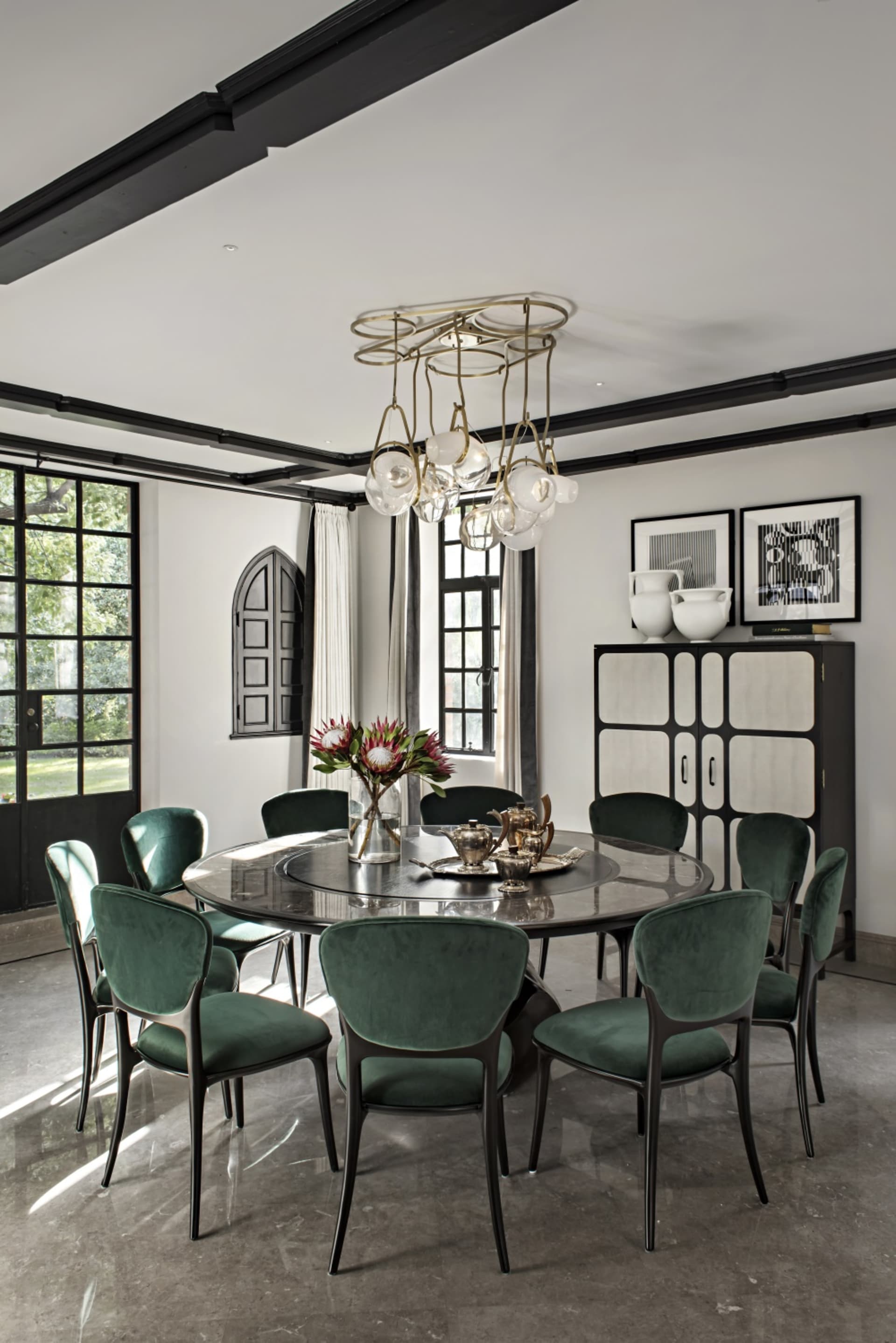 A modern dining room featuring a round table with marble top, surrounded by green velvet chairs with black frames, and a hang