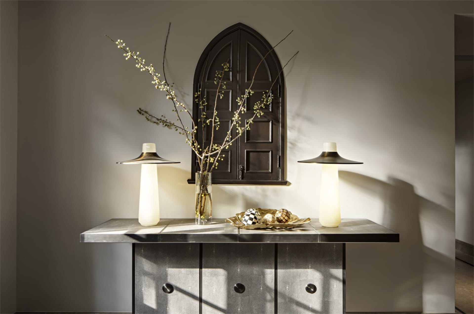 A ceramic decorative table with a vase of flowers, two lamps, and decorative objects on top of it, against a wall with a wind