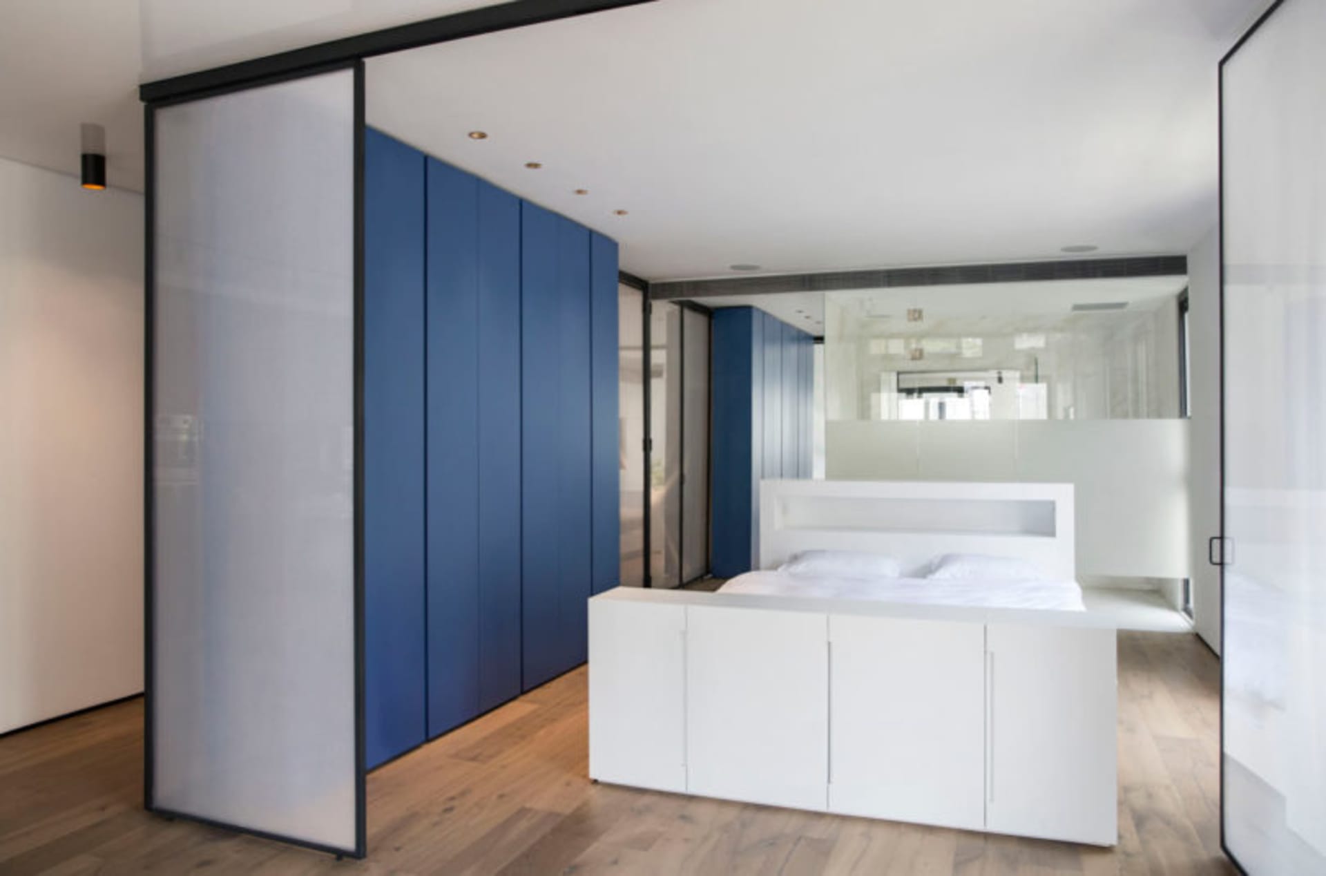 Modern bedroom with white furniture, blue storage cabinets, and glass partition, featuring white floor and wood flooring.