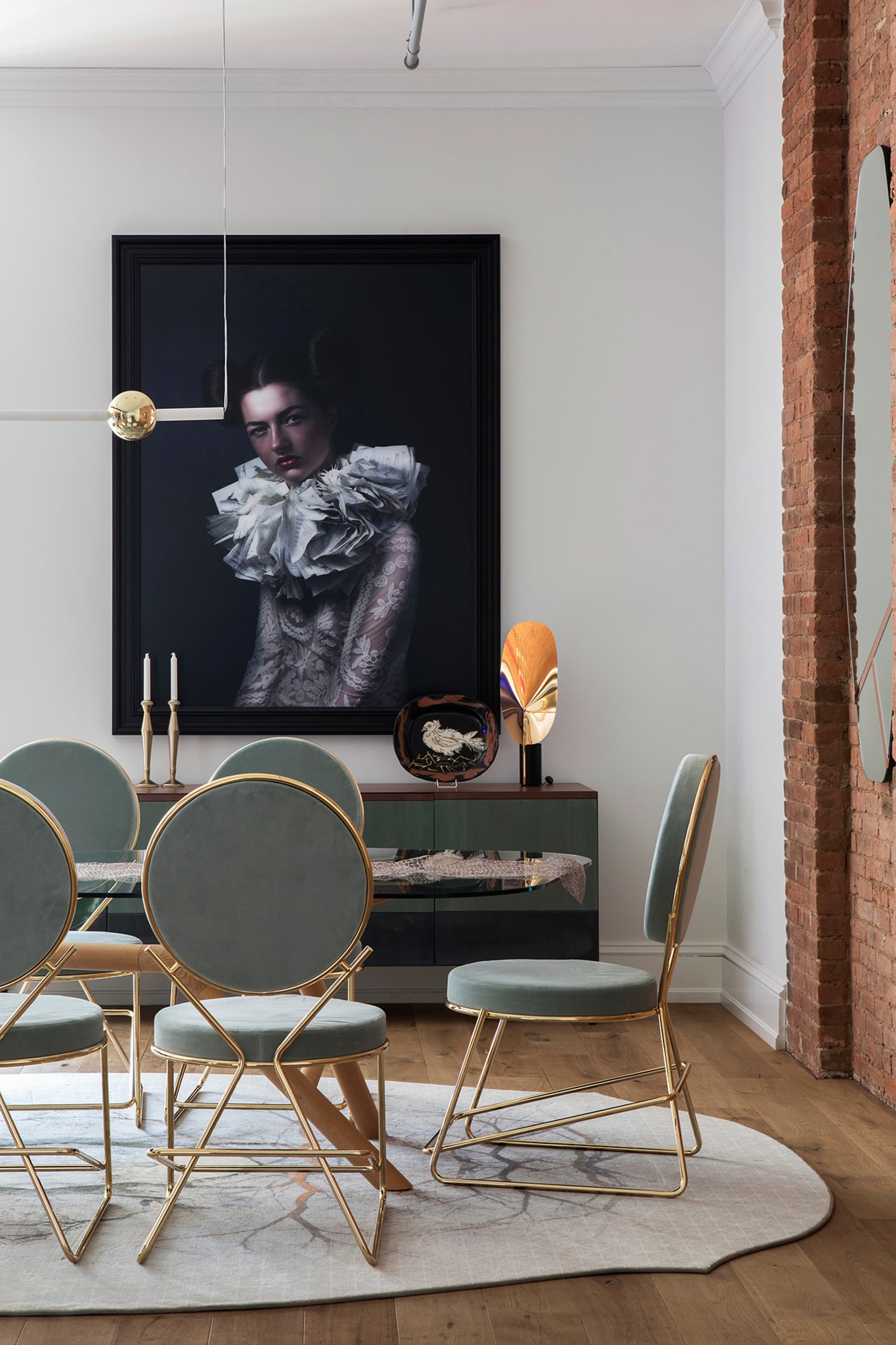 A modern dining room features metallic chairs with blue velvet upholstery, a glass table, and a large portrait of a woman.