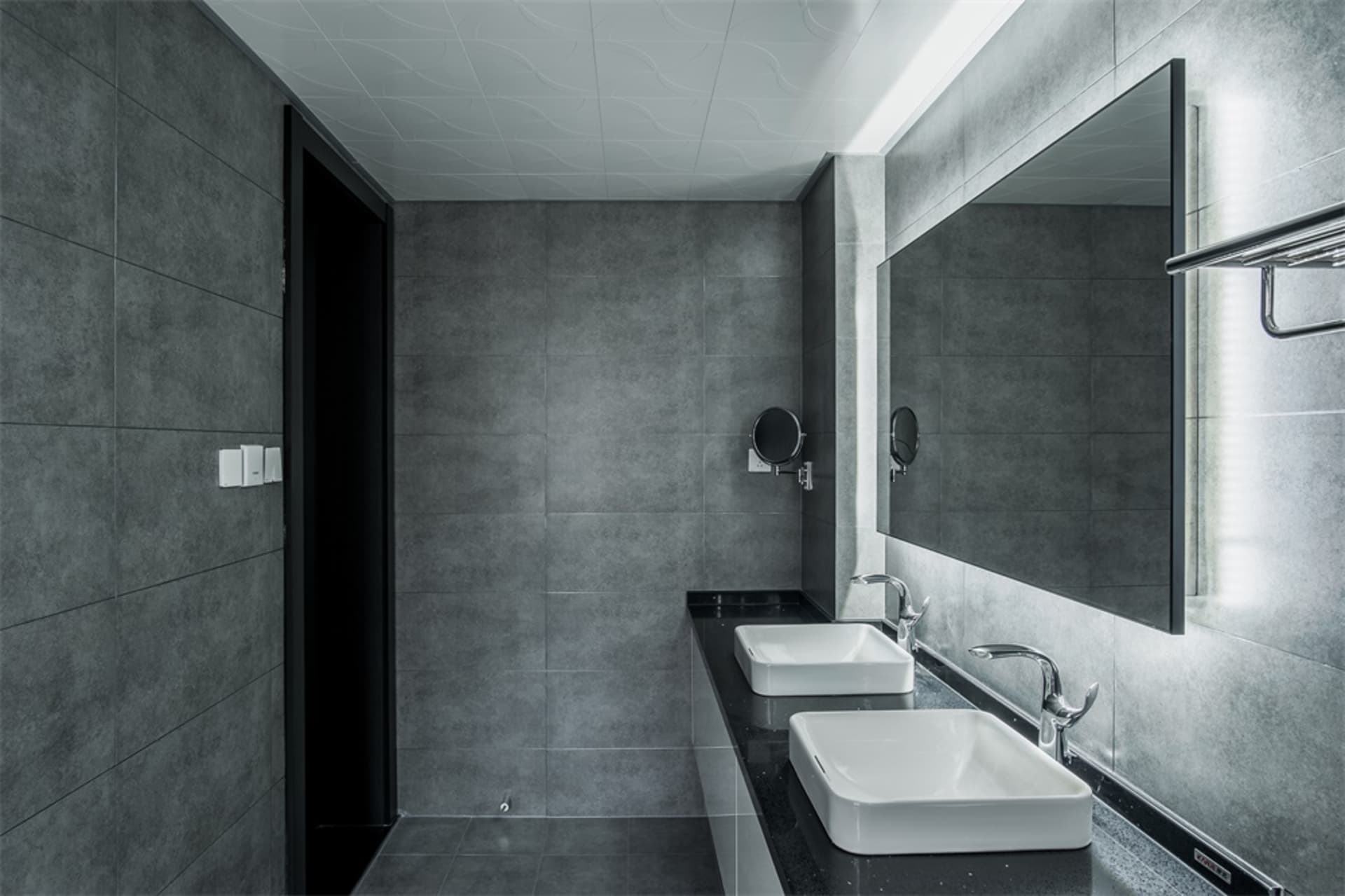 A modern bathroom with black and gray ceramic tiles, a white countertop sink, and a large rectangular mirror.