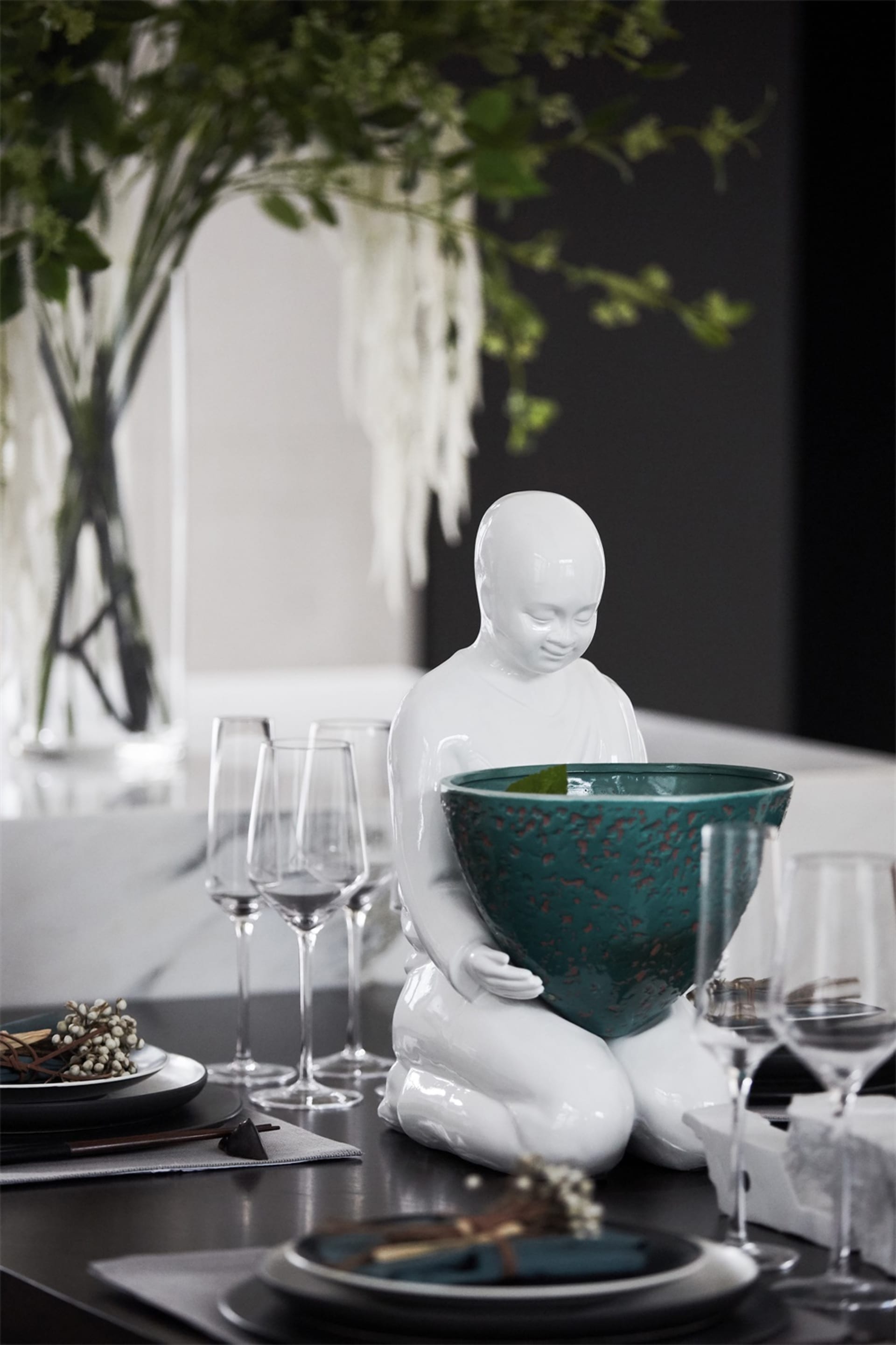 A modern interior design table setting with a white ceramic statue holding a green bowl, surrounded by wine glasses and plate