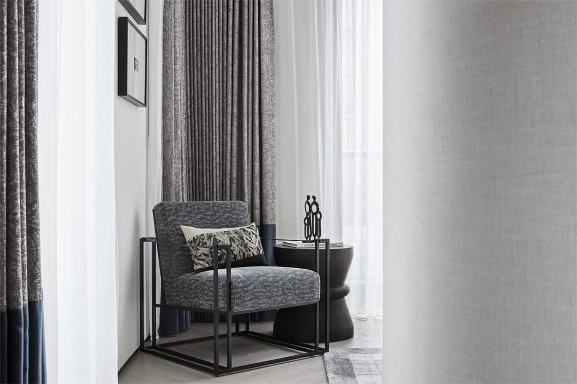 A modern minimalist living room featuring a grey upholstered chair with metal frame, white curtains, and a black side table.