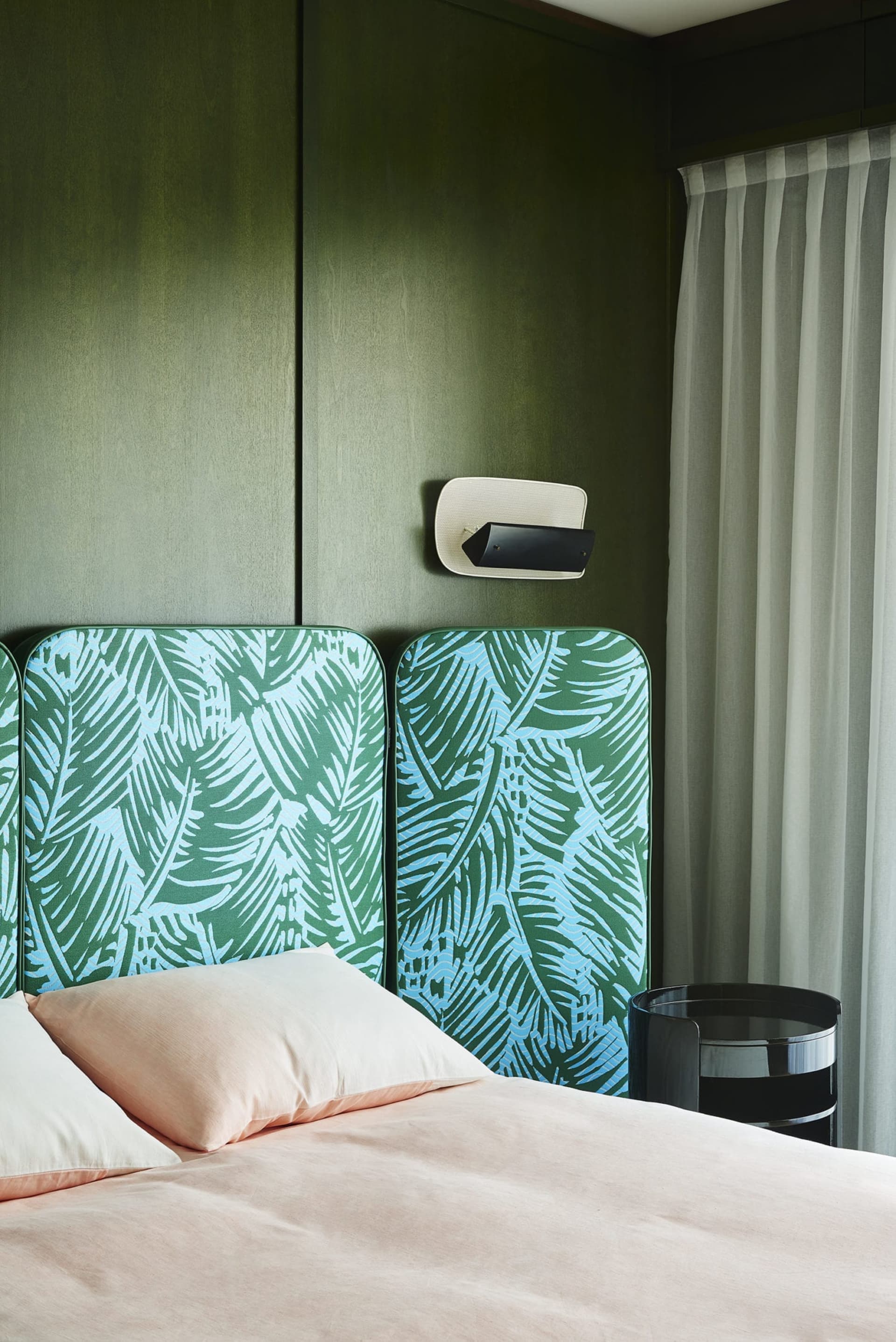A modern bedroom featuring a bed with pink bedding, a green palm leaf print headboard, and a window with sheer curtains.