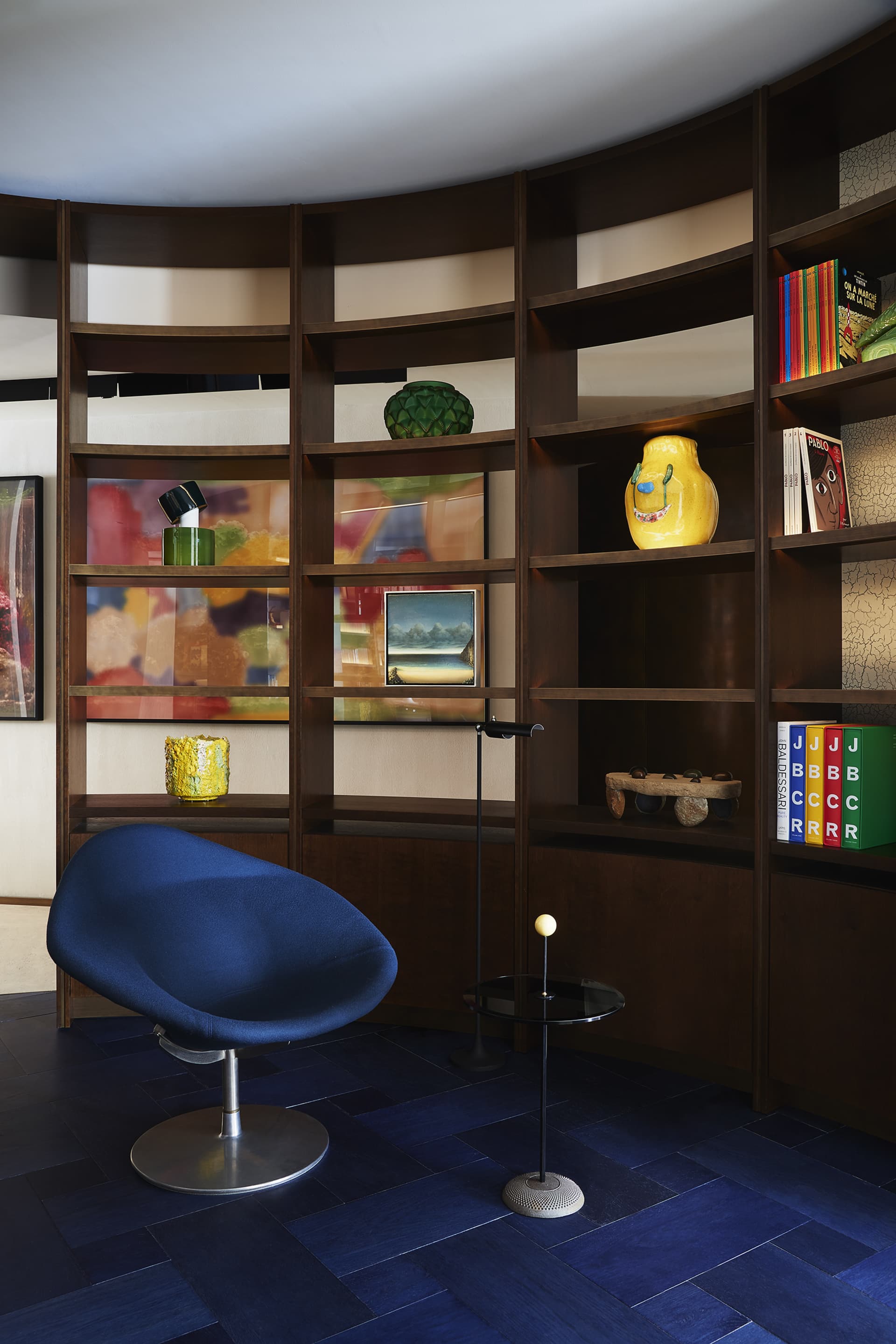 A modern home office featuring a curved wooden bookshelf filled with colorful objects and books.