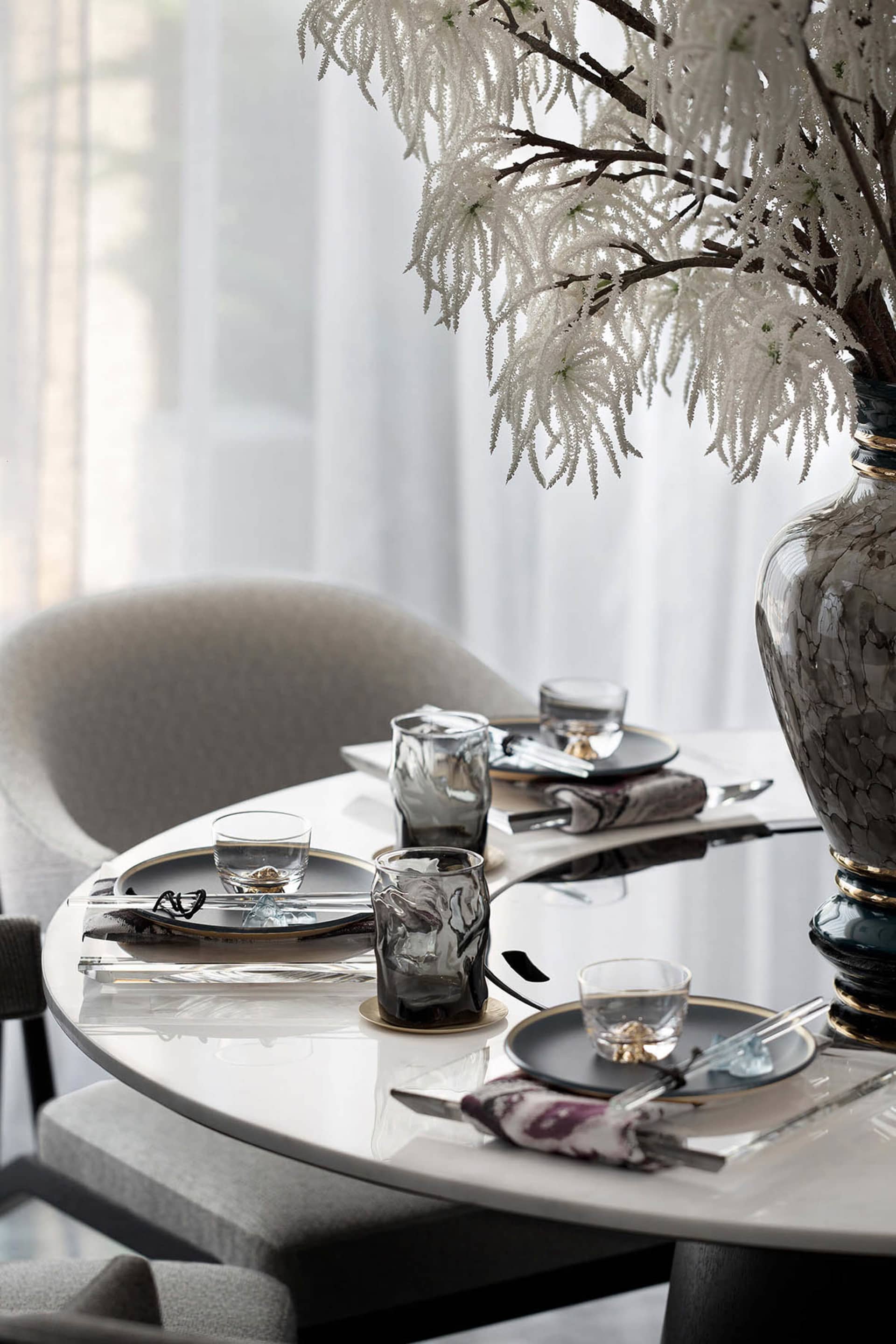 Modern dining room featuring sleek glass tables and chairs, with white curtains and decorative white flowers in a vase.