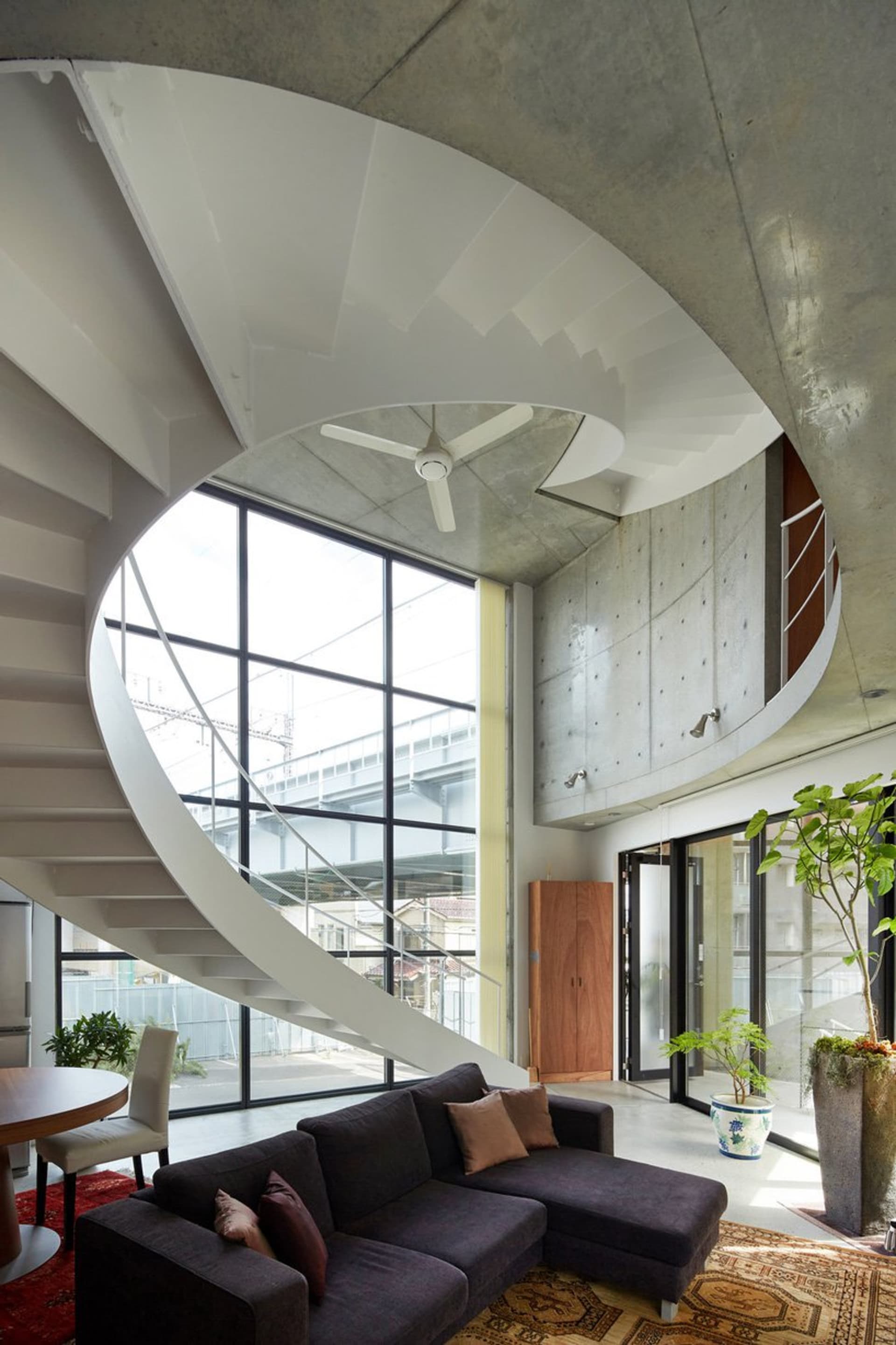 Modern residential living room with a spiral concrete staircase, large windows, and a dark sofa.