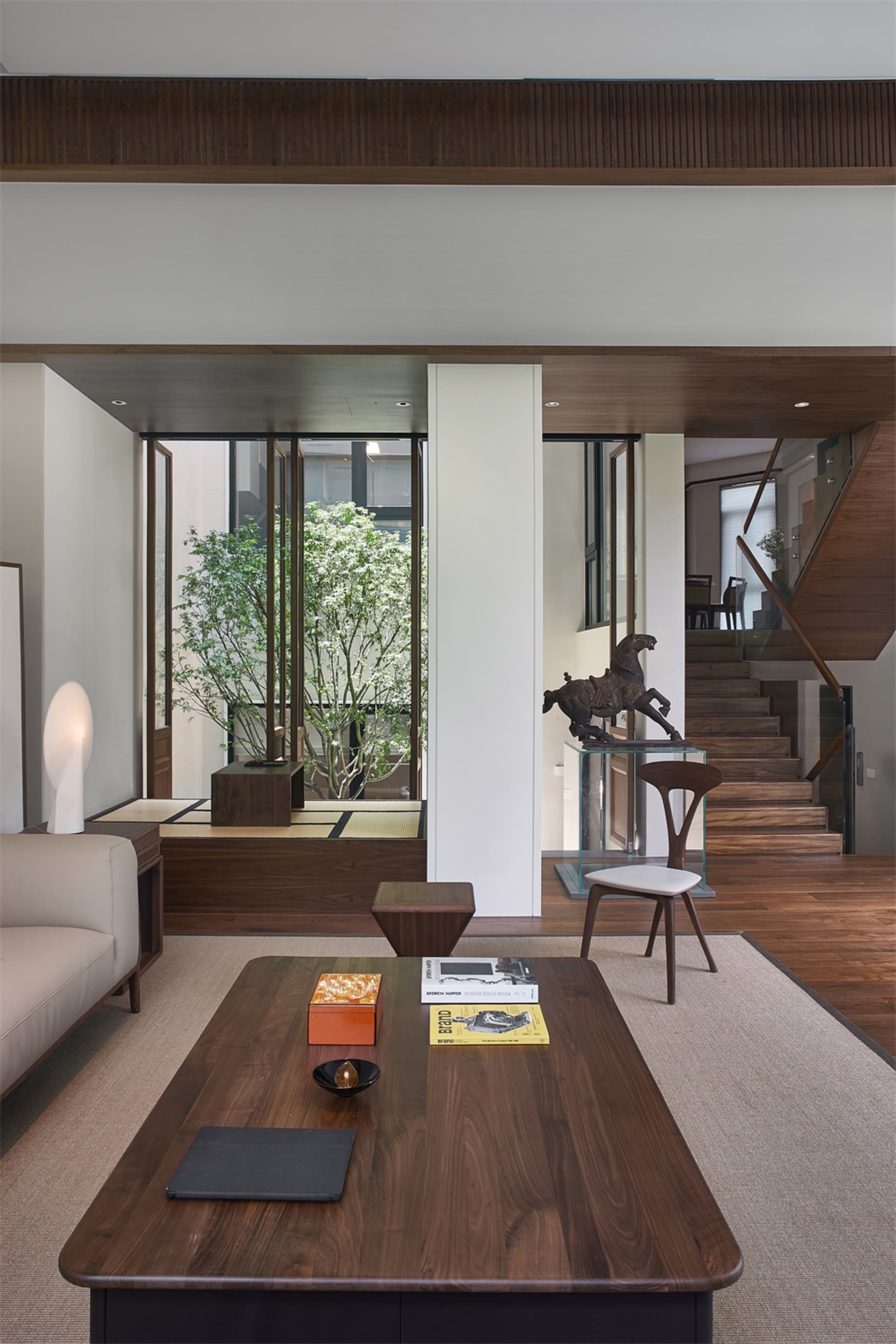 A modern living room with wooden furniture, featuring a large window, a coffee table with books and a bowl, and a wooden floo