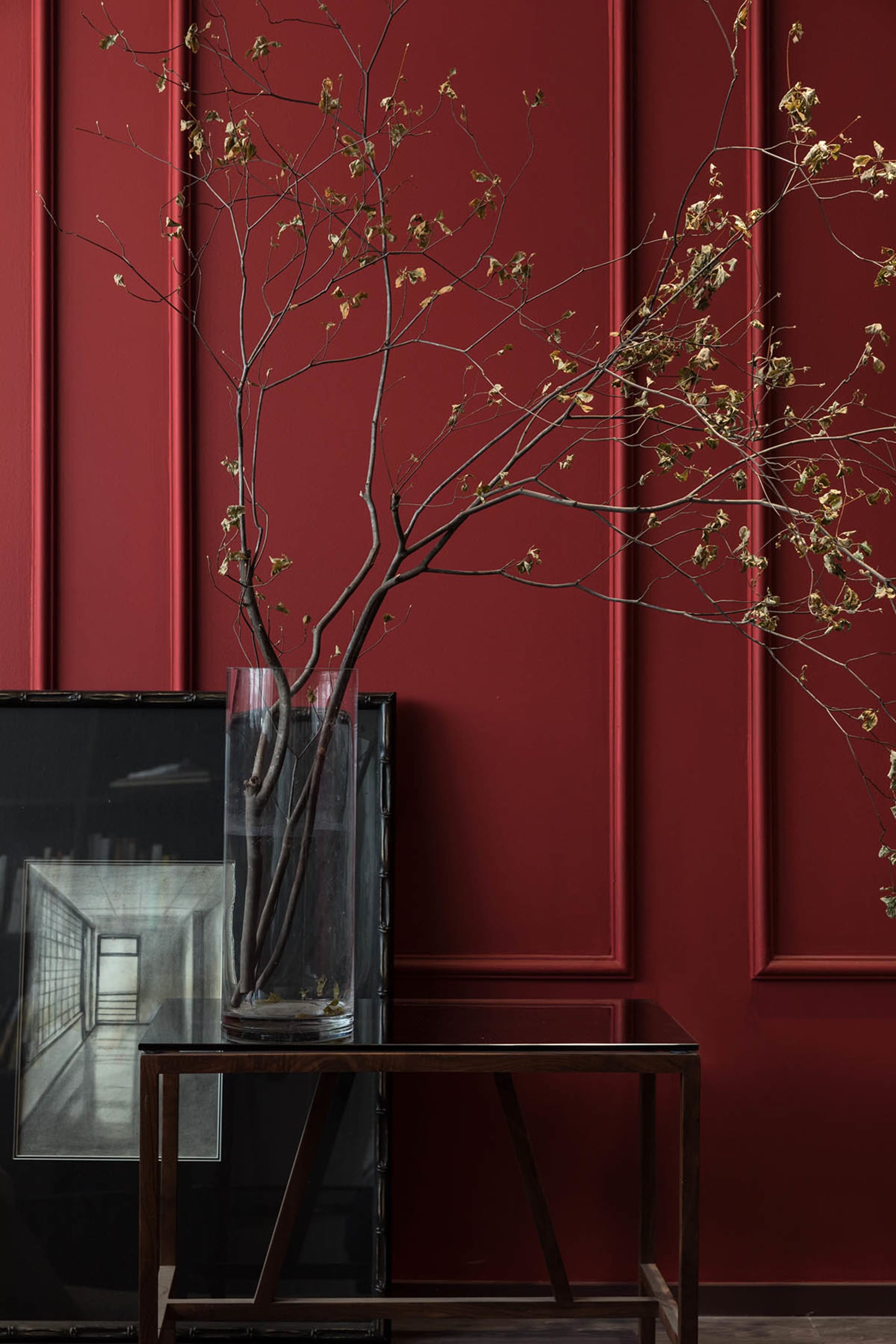A vase containing branches with dried leaves on a wooden side table in a red-walled room.