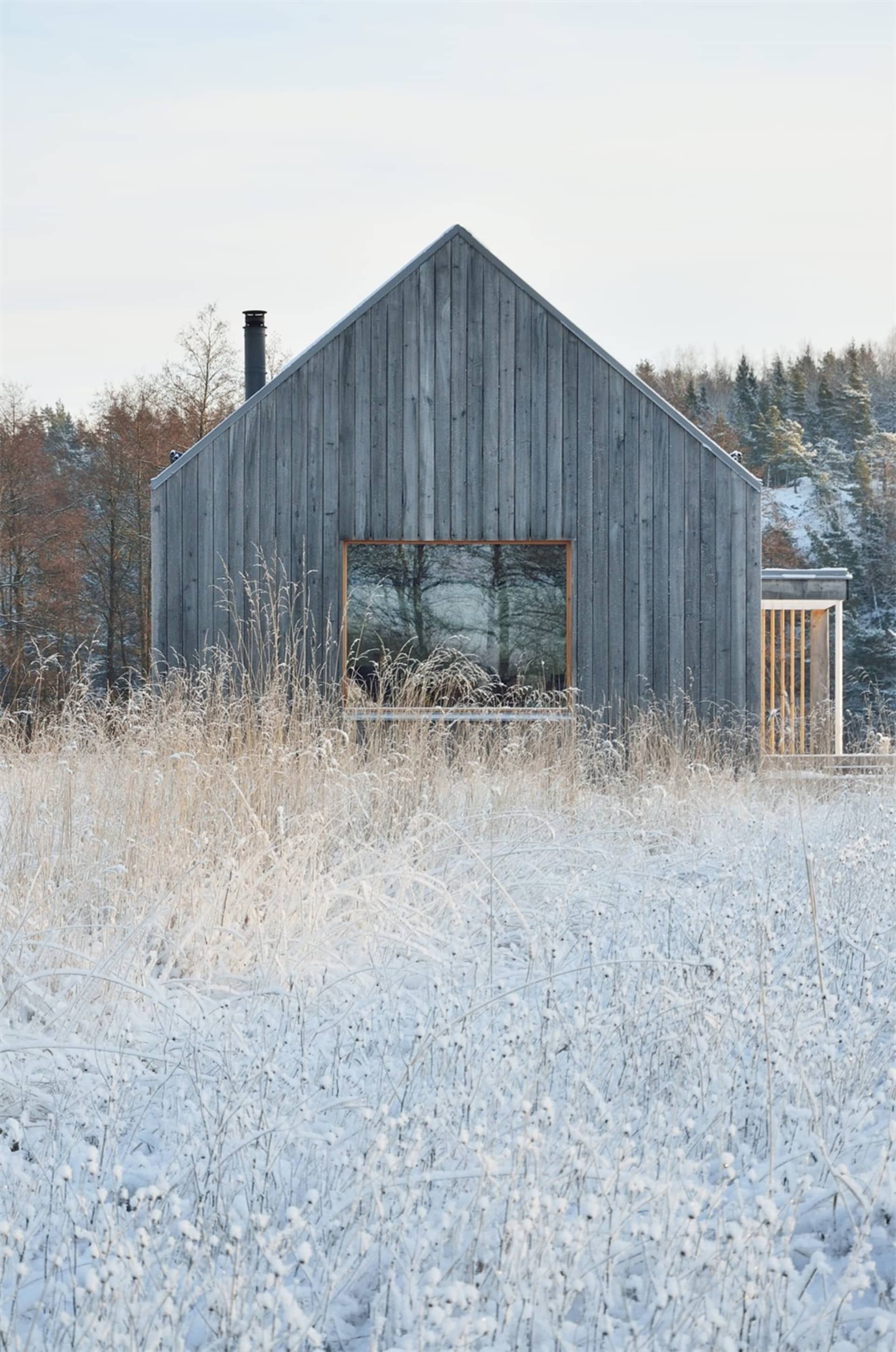 A minimalist wooden house surrounded by snow-covered grass and trees in a winter landscape.