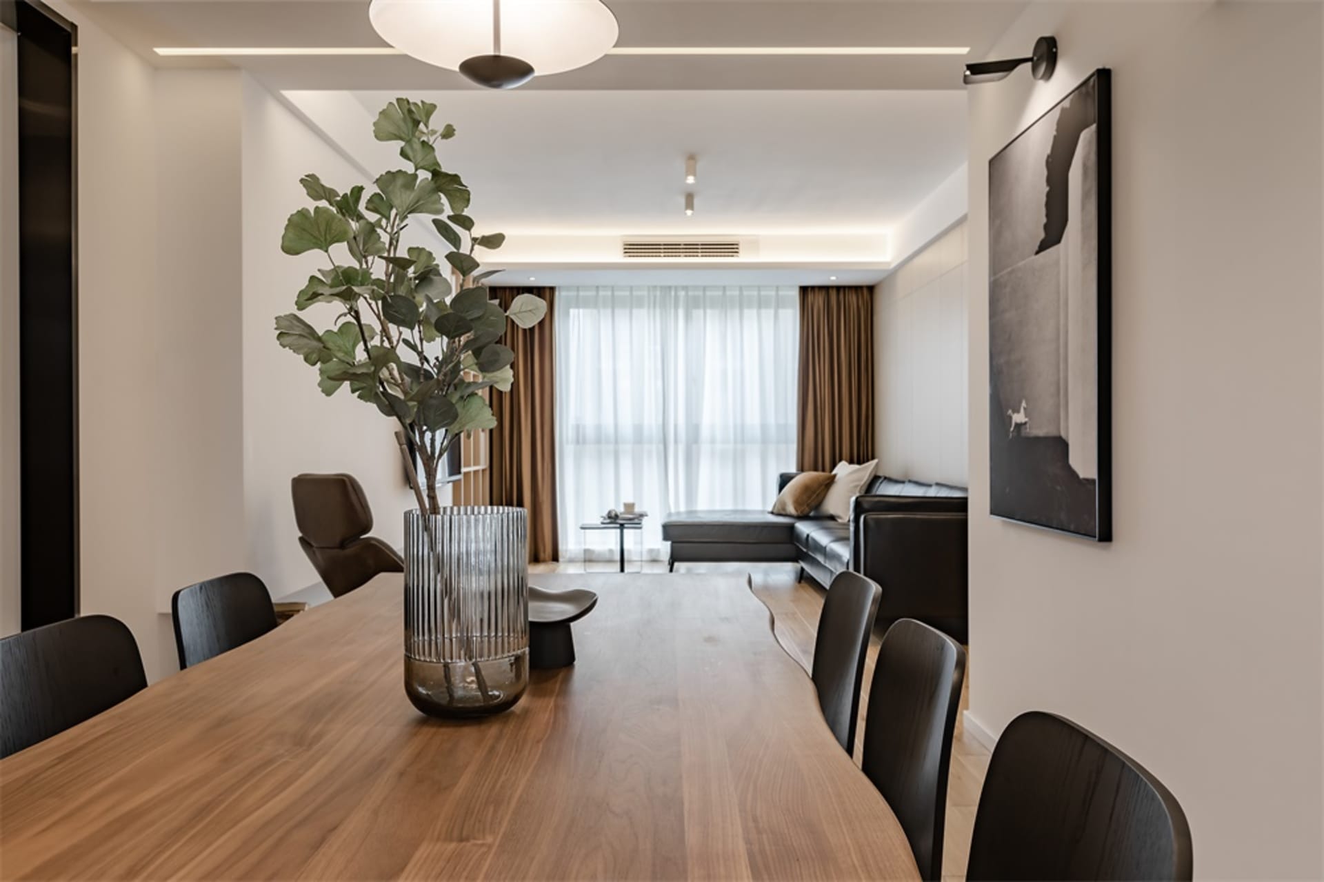 A modern living room featuring a wooden table with a green plant in a clear vase, black chairs, a gray leather sofa, and a la