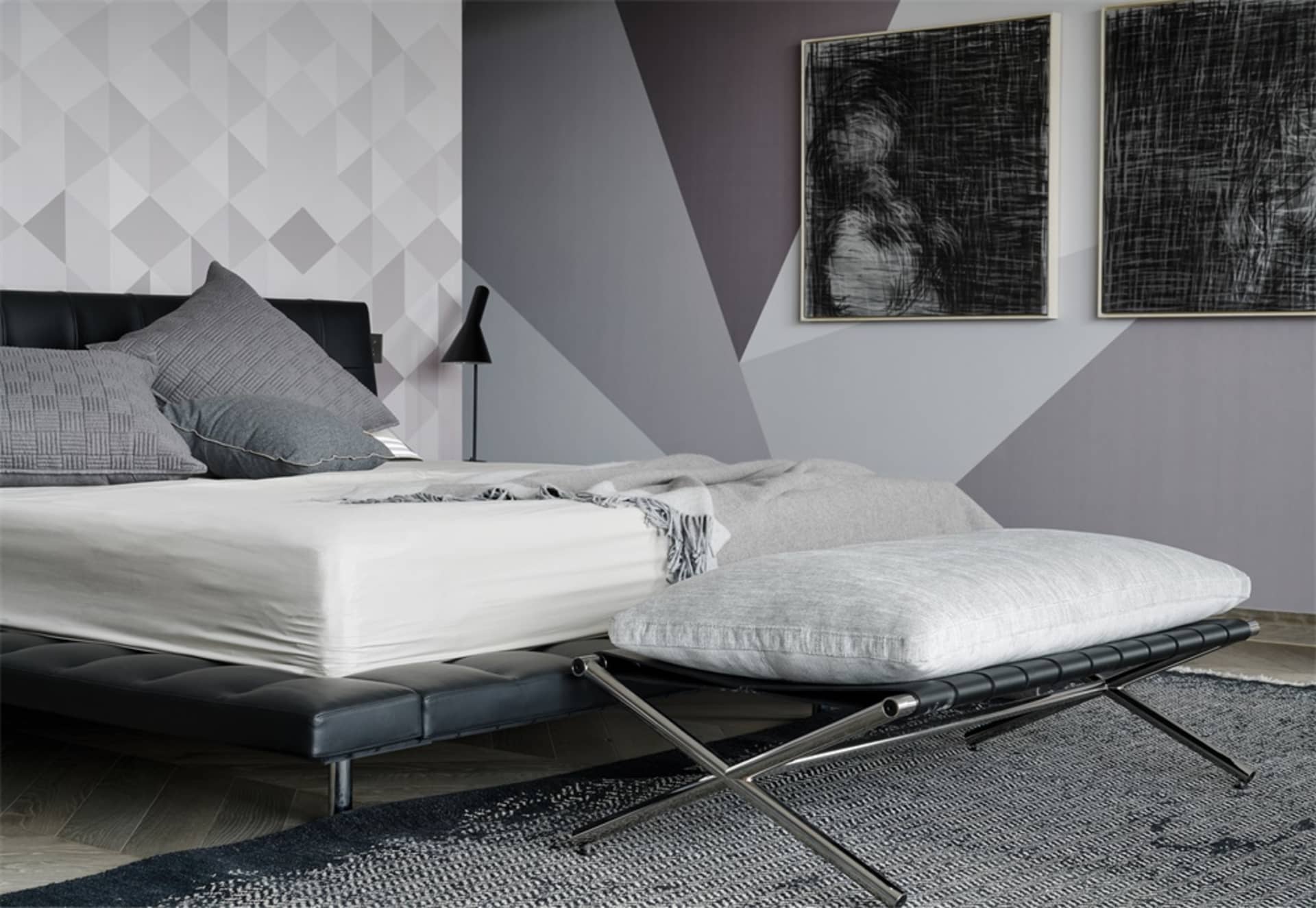A modern minimalist bedroom featuring a black leather bed with grey pillows, a white blanket, and a grey upholstered bench.