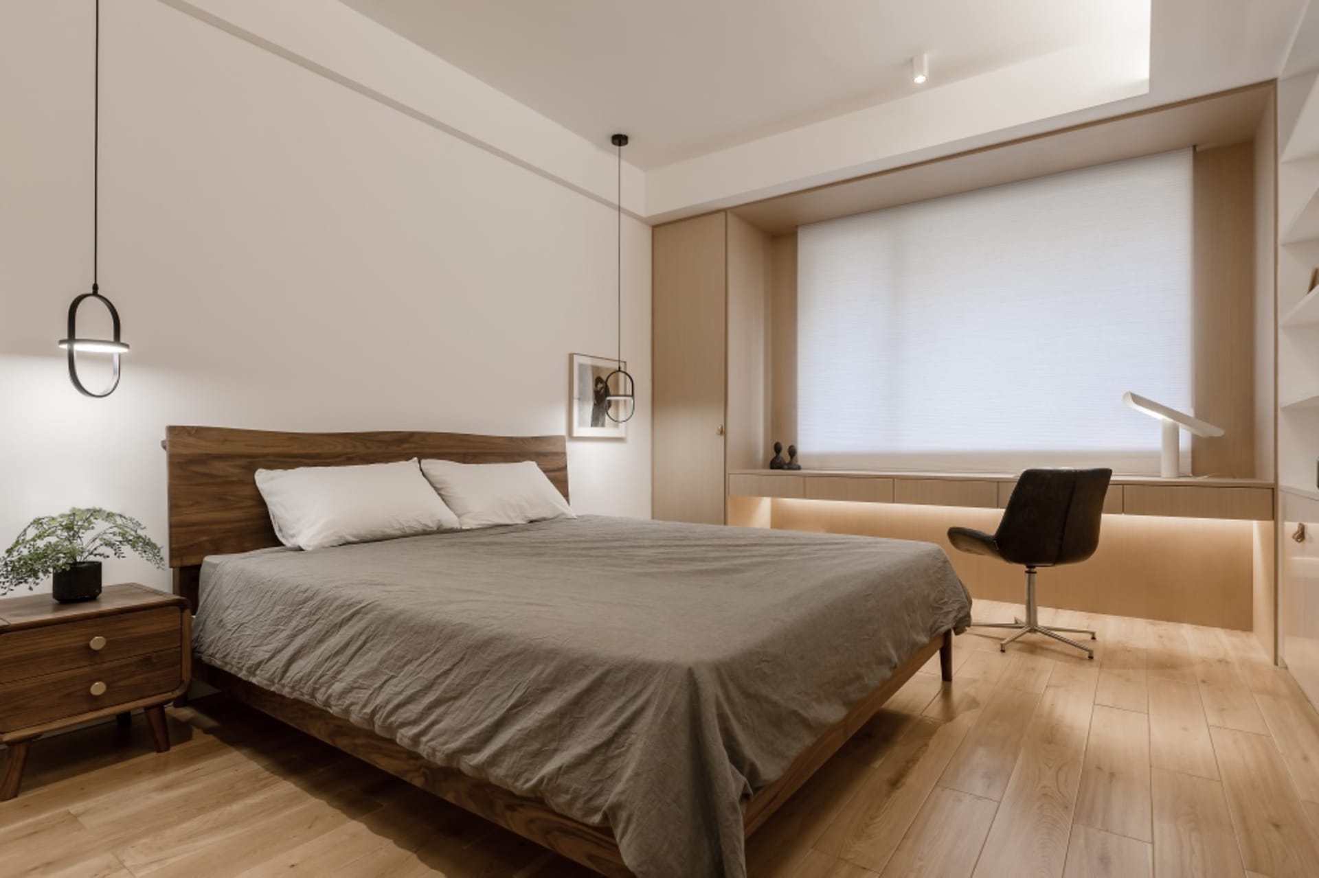 A minimalist bedroom with a wooden bed and a black chair, featuring a large window with white blinds and wood flooring.