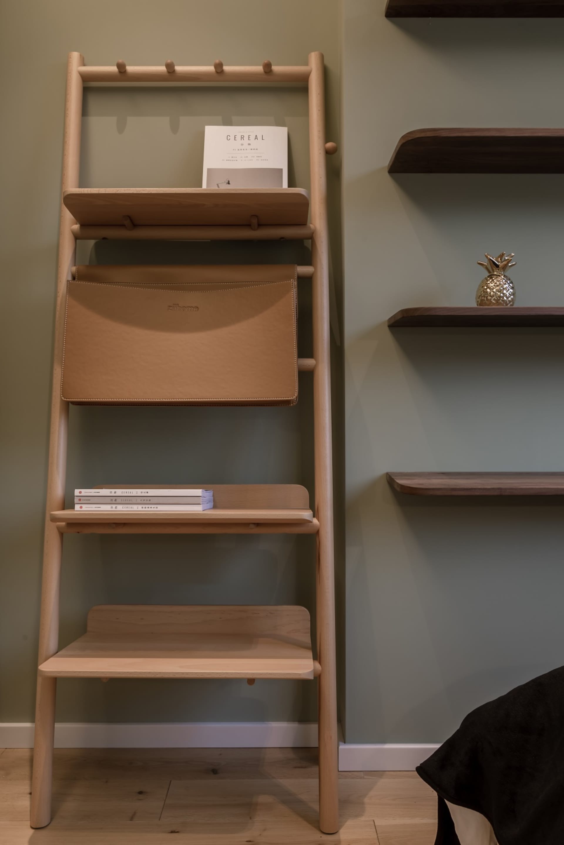 A wooden bookshelf with shelves and a bag attached to it, featuring a book and a golden pineapple ornament.