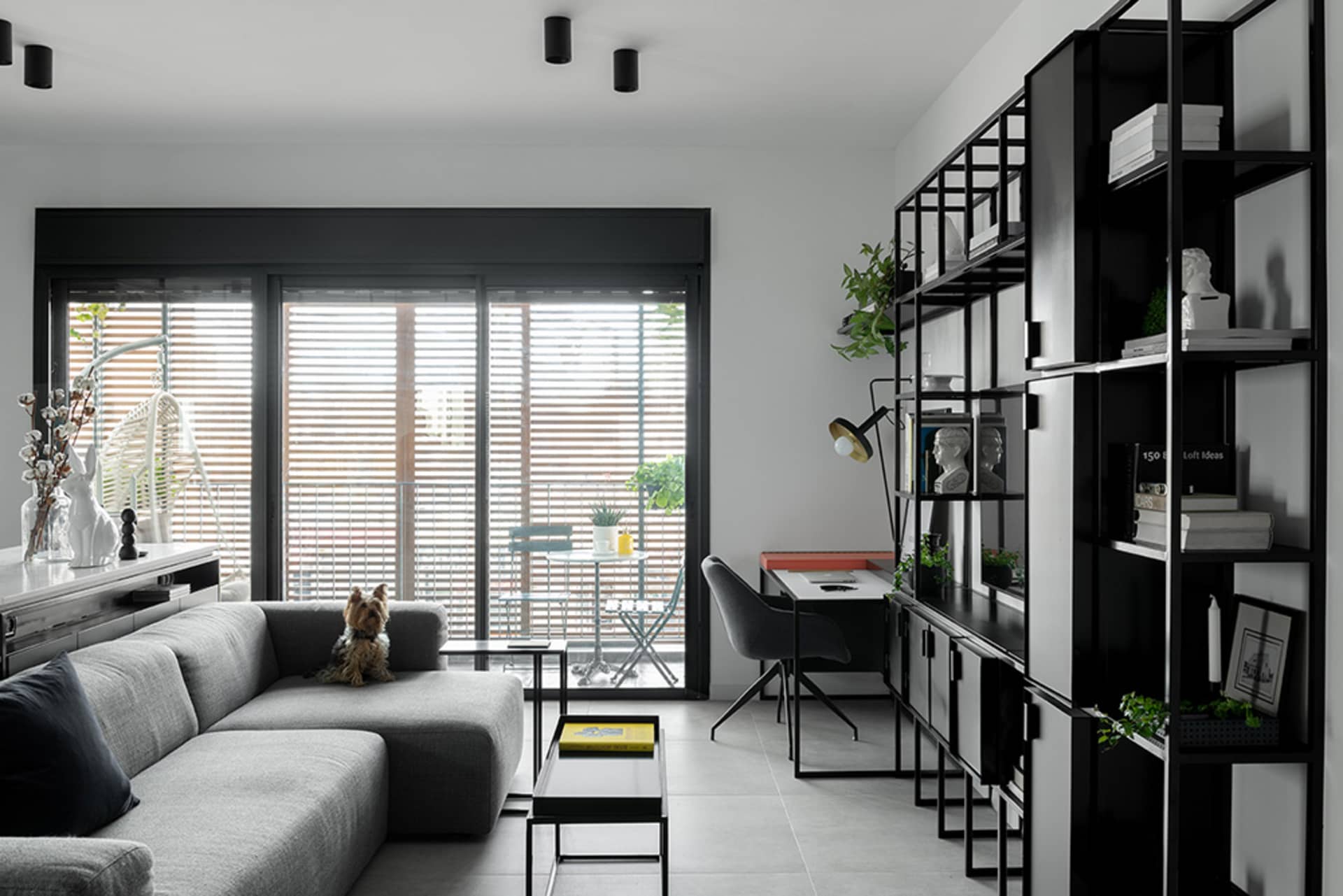 A modern minimalist living room featuring a grey sectional sofa, a glass coffee table, and a black and white shelving unit wi