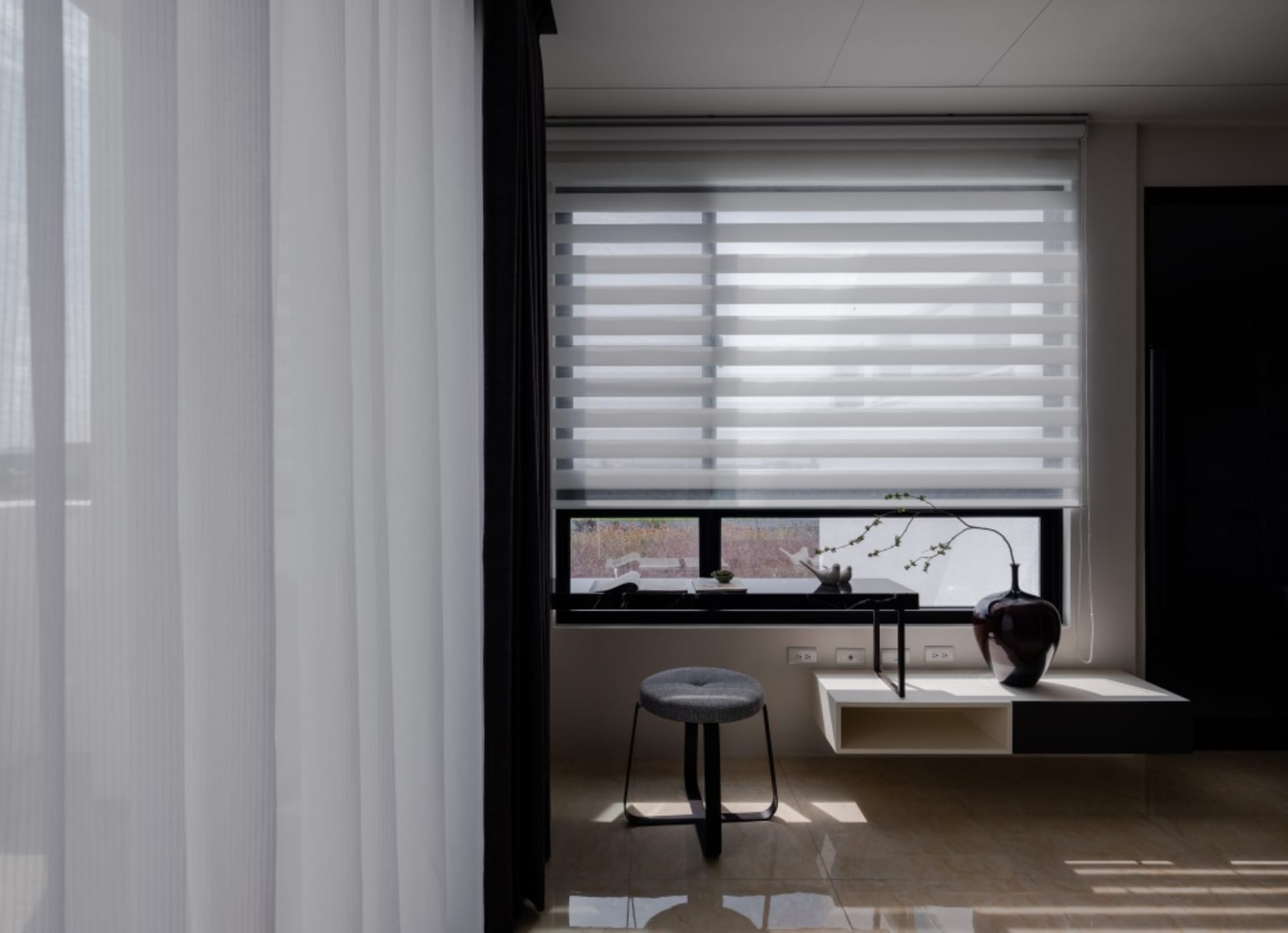 A modern living space featuring minimalist window blinds, a wooden stool, and a vase with a plant.