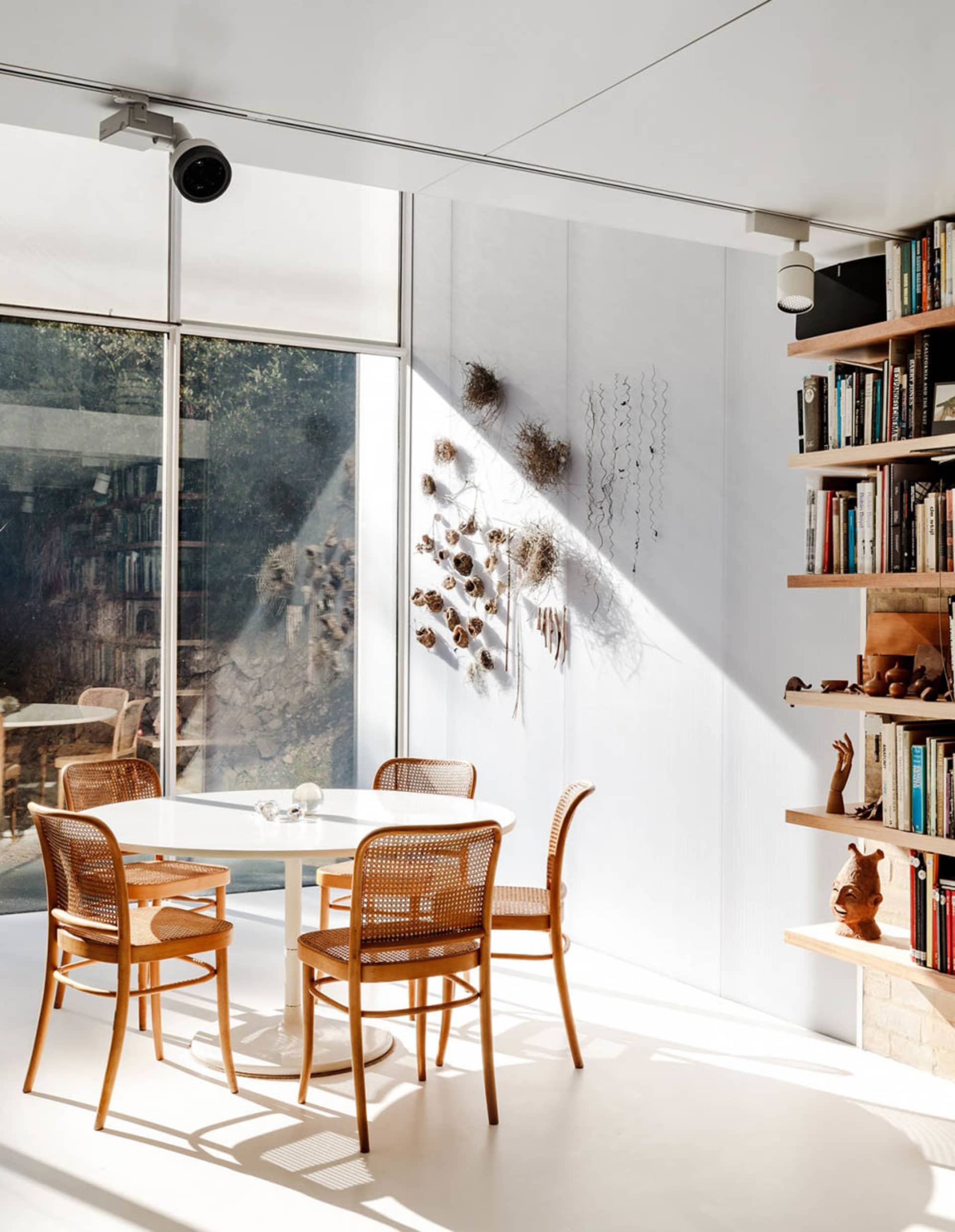 A modern dining room with a white table, wooden chairs, and glass wall providing a view of the outdoors.