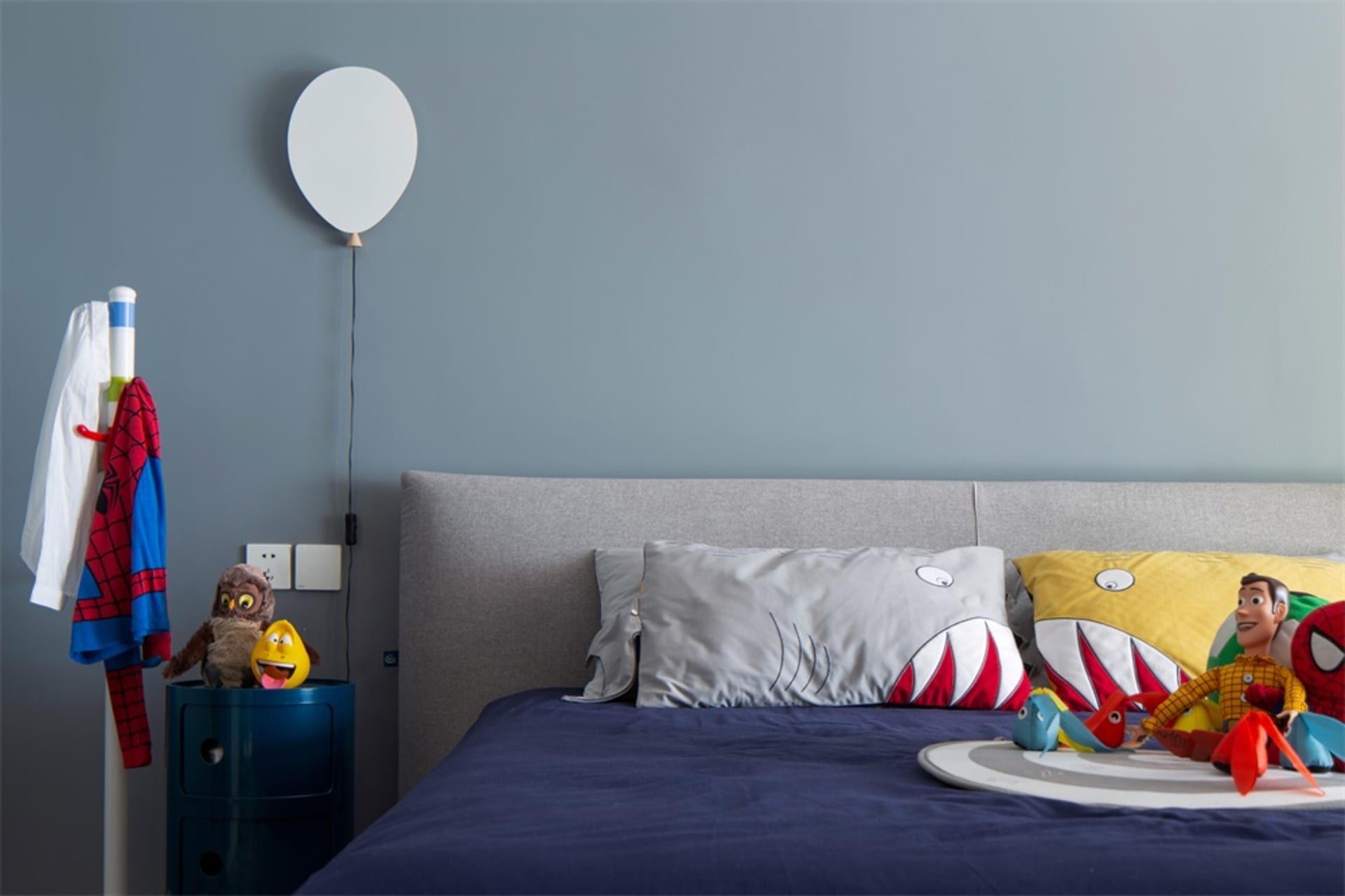 A modern bedroom with a gray upholstered headboard, blue bedding, and colorful plush toys.
