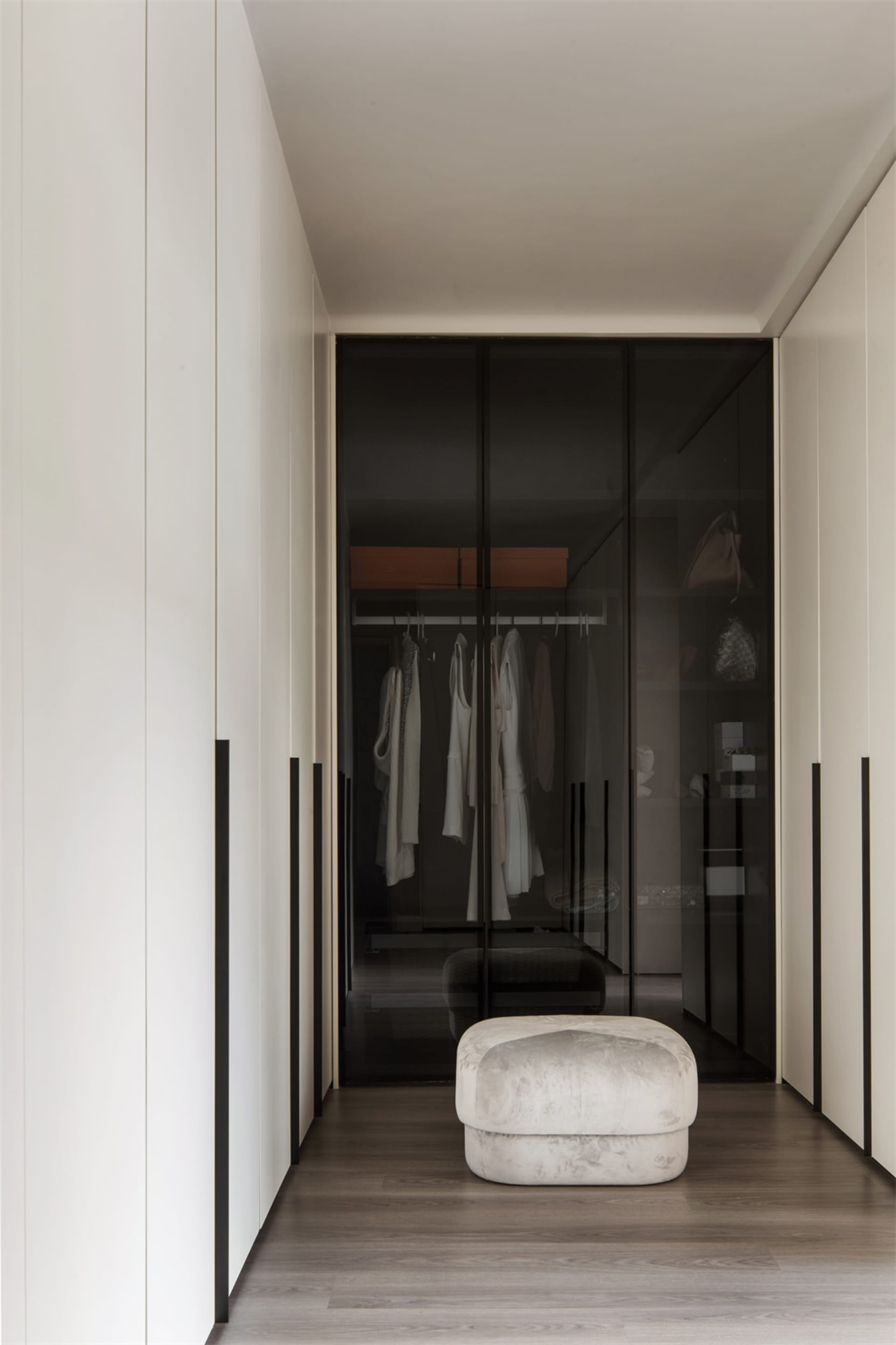 An image of a modern minimalist closet featuring glass sheets and a cushioned ottoman on a wooden floor.