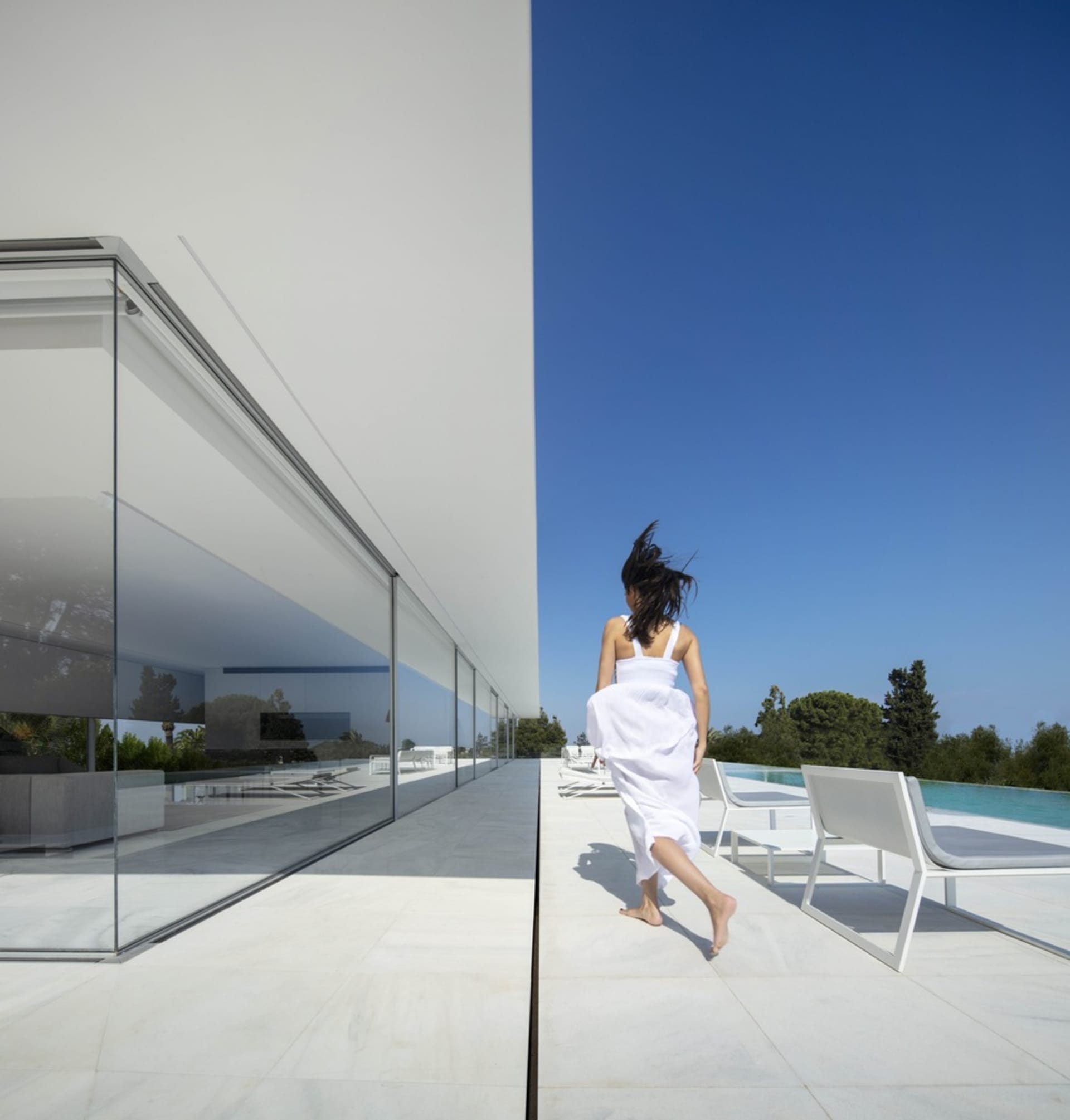 A woman in a white dress walking on a patio with white concrete flooring and a modern house exterior featuring glass walls an