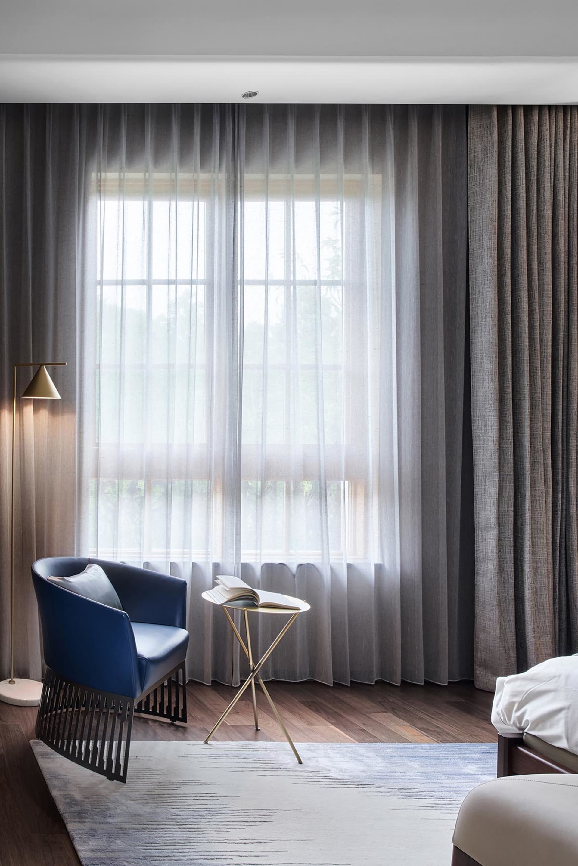 A modern bedroom with a white bed, blue chair, and textured cotton curtains behind it.
