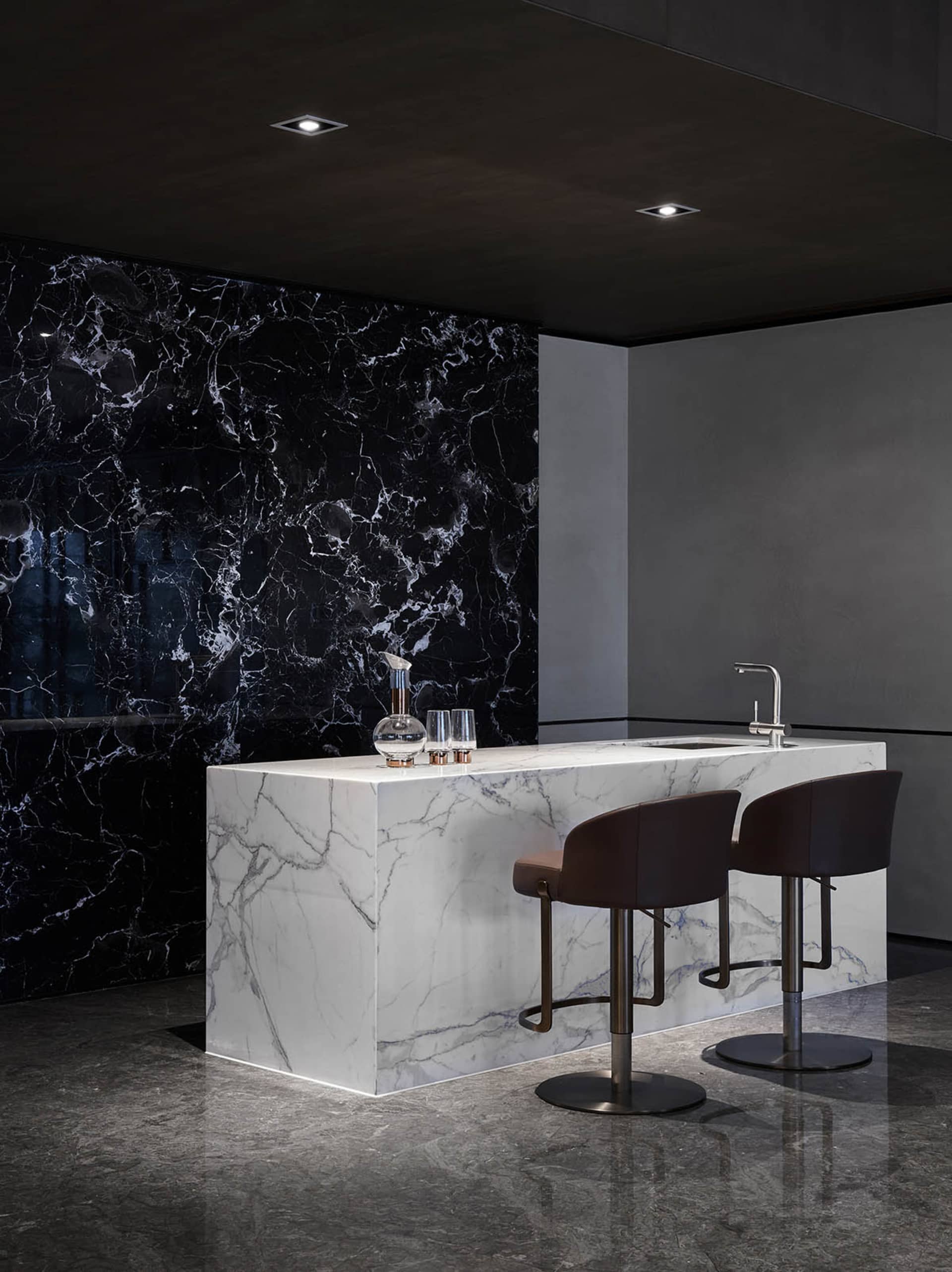Modern kitchen with white marble countertop and black marble backsplash, featuring a single water faucet and two bar stools.