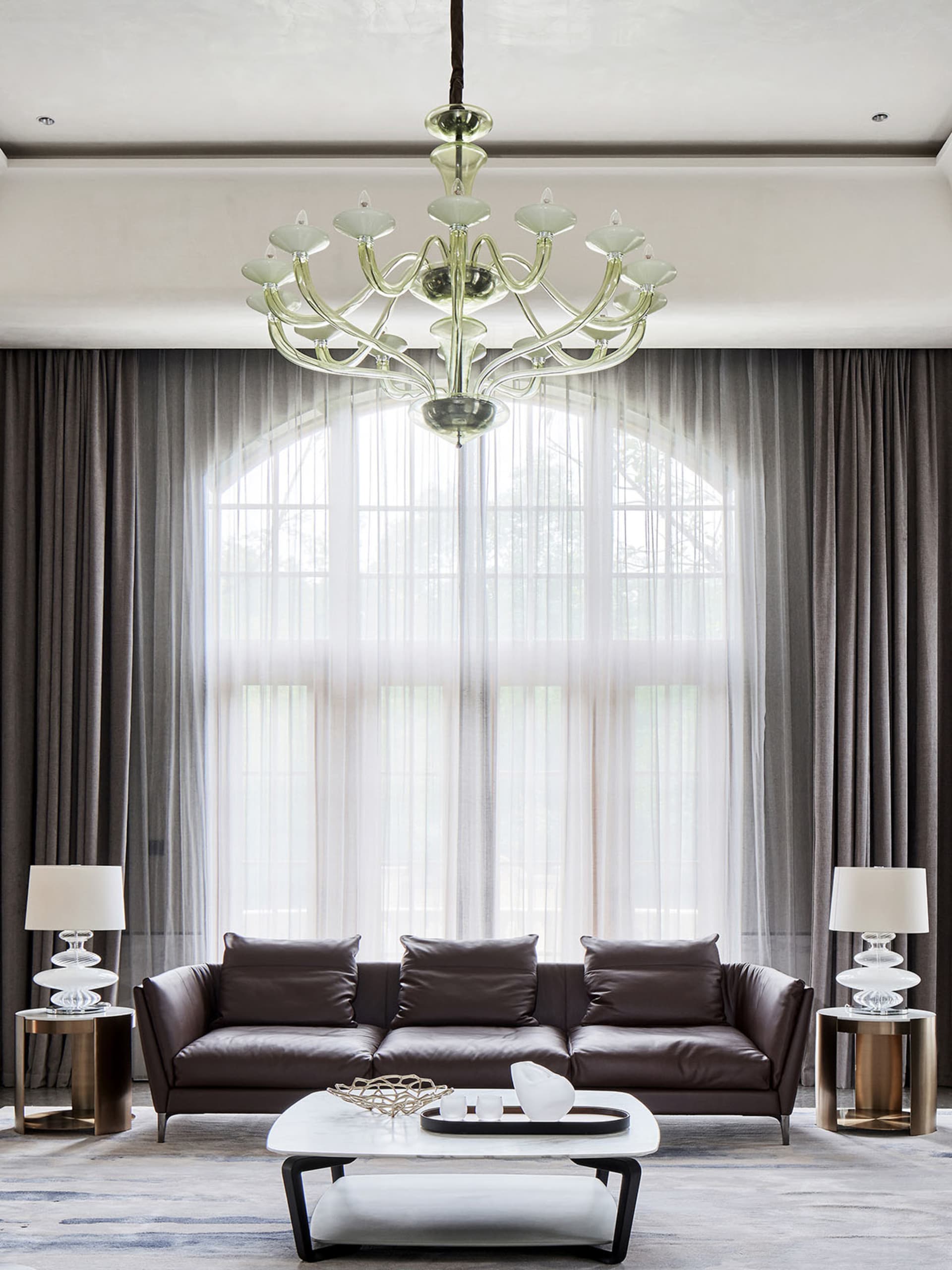 A modern living room with a dark leather sofa, glass coffee table, and metal side tables. The room features large windows wit