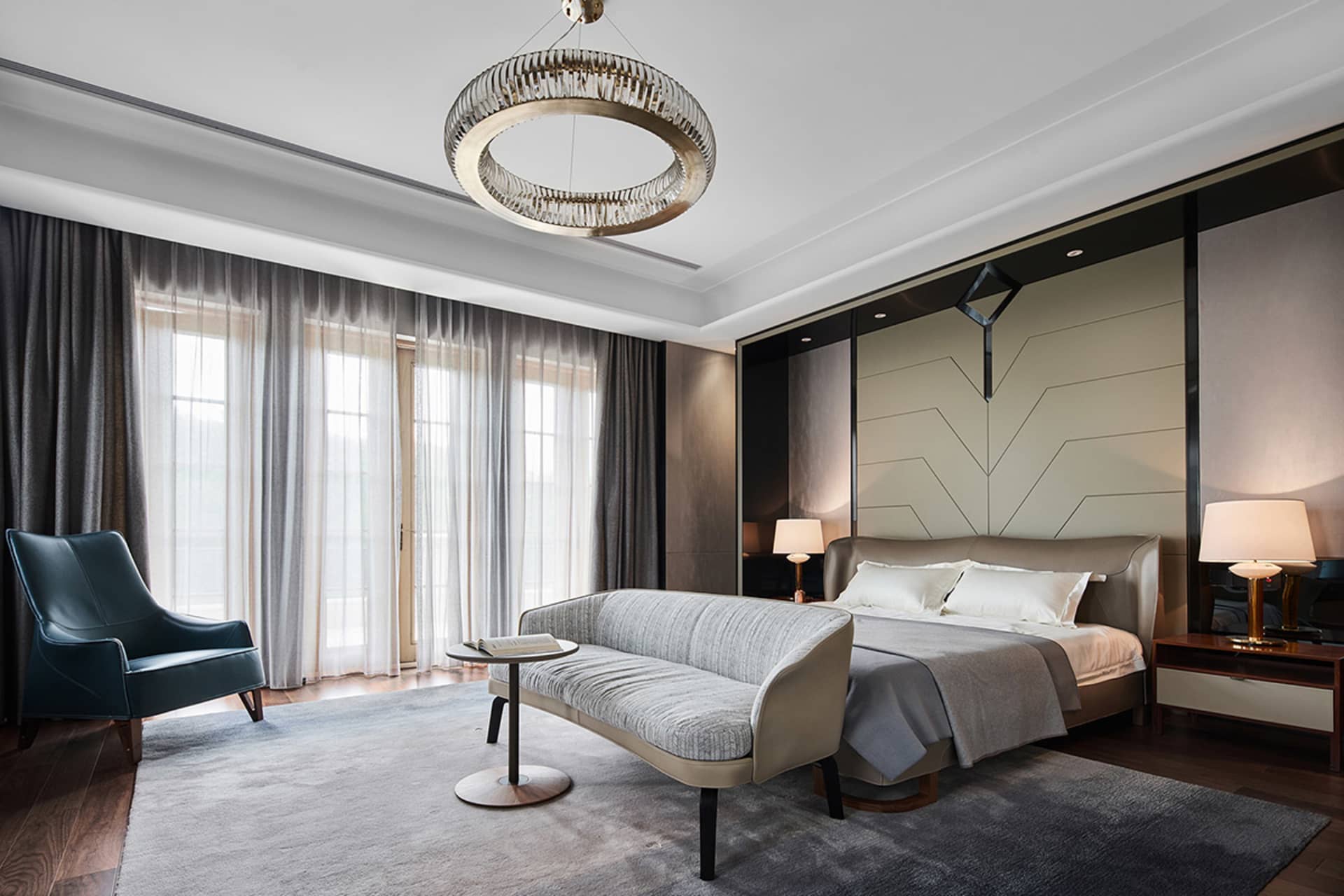 A modern residential bedroom with grey fabric furniture, wooden accents, and a sleek glass lighting fixture.