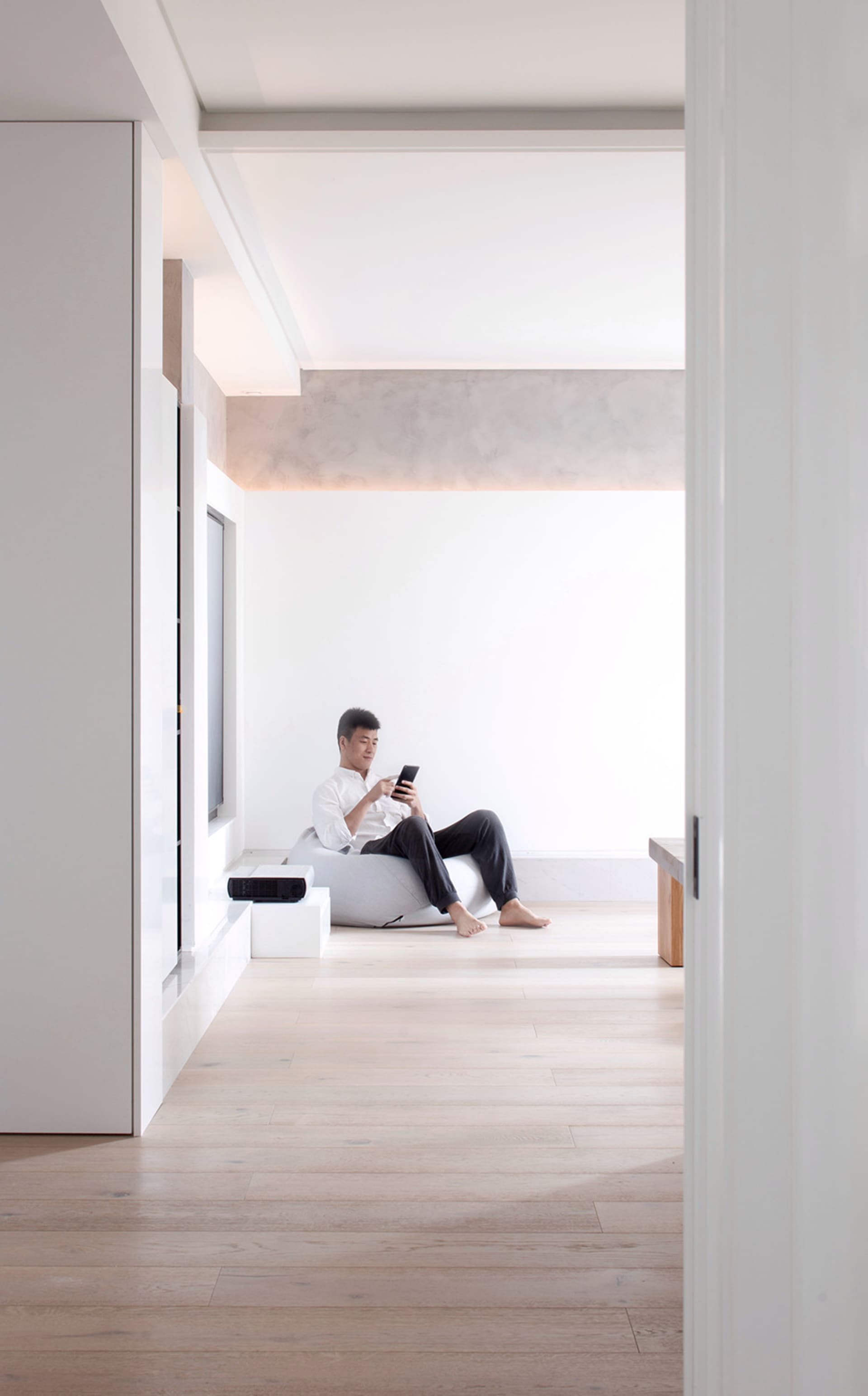 Person sitting on a beige fabric ottoman in a minimalist living room, looking at a smartphone, floor-to-ceiling white walls,