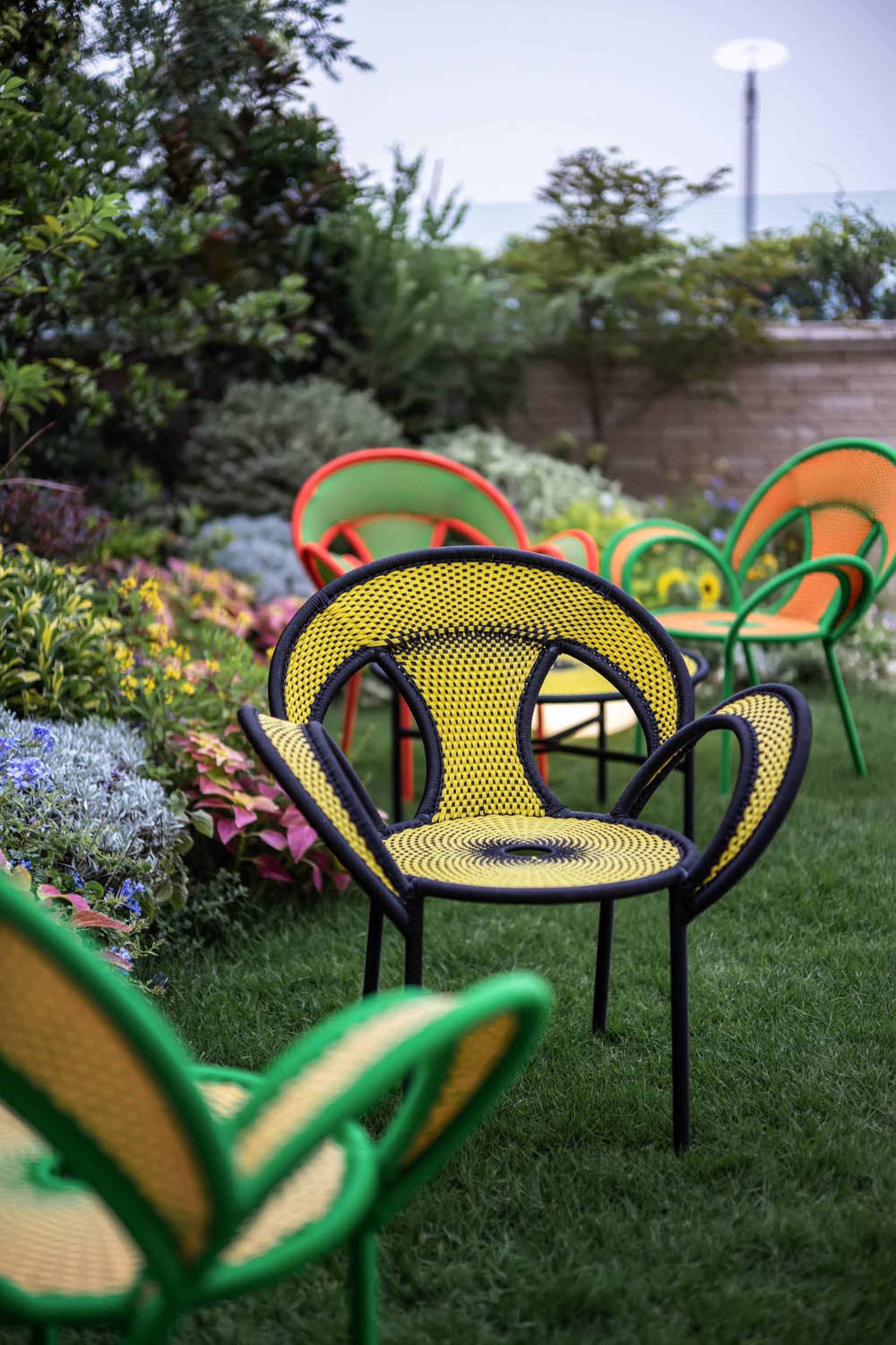 A modern, yellow, geometric-patterned garden chair made of metal and woven fabric surrounded by colorful flowers and plants.