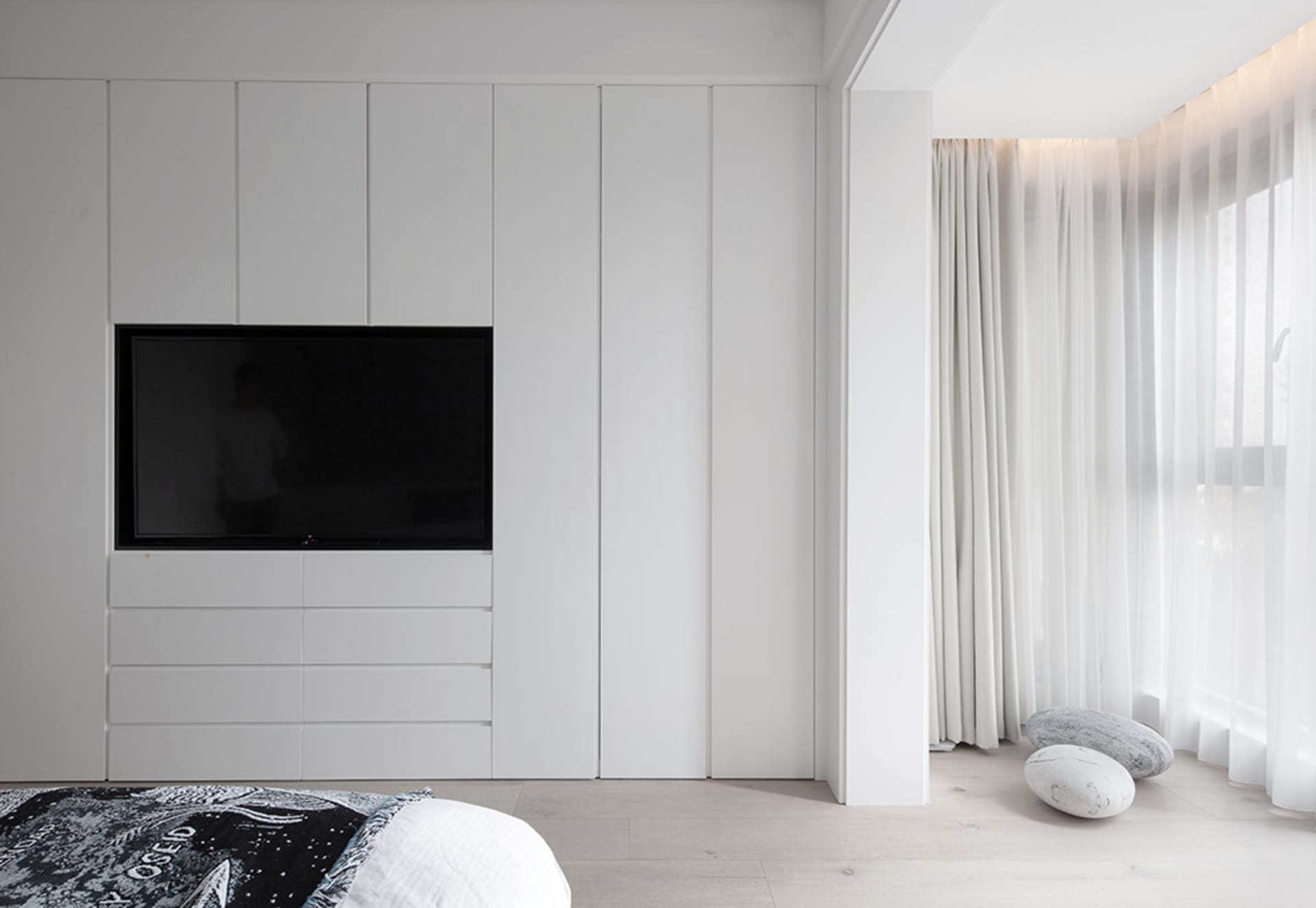 A minimalist bedroom with white furniture and a television on the wall.
