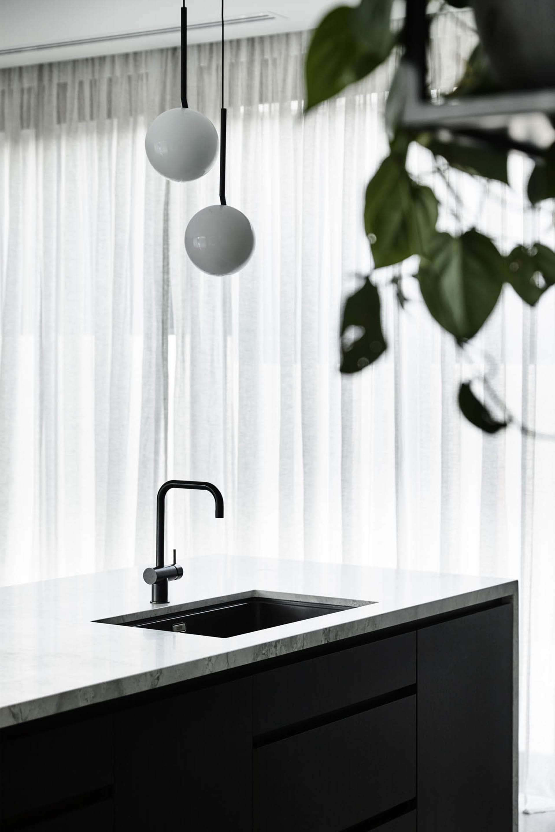 Modern kitchen with black and white marble countertops, black sink, and hanging white globe lights. White sheer curtains and