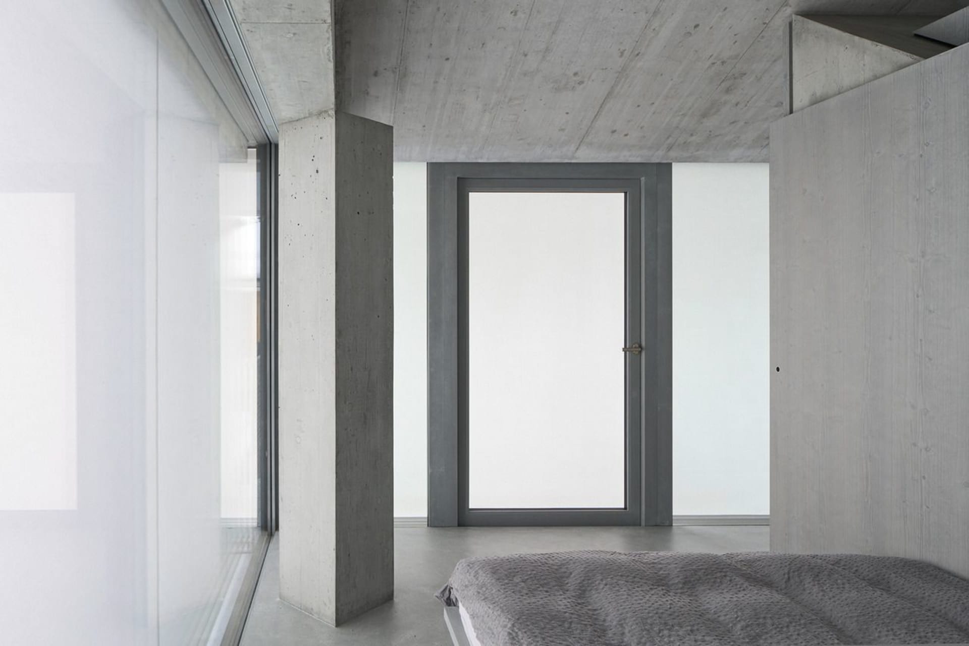 A minimalist bedroom with concrete walls, floor, and ceiling, featuring a bed with a gray cover.