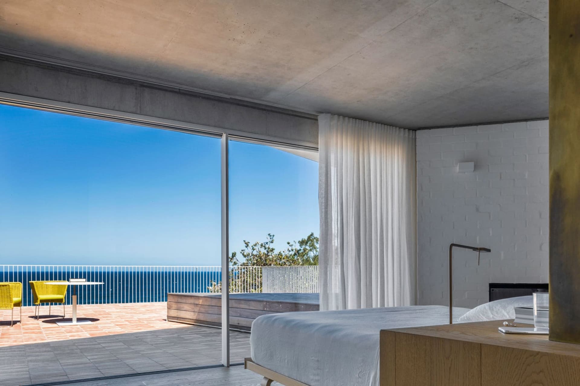 Modern residential bedroom featuring a minimalist design with a light concrete floor, white sheer curtains, and a view of the