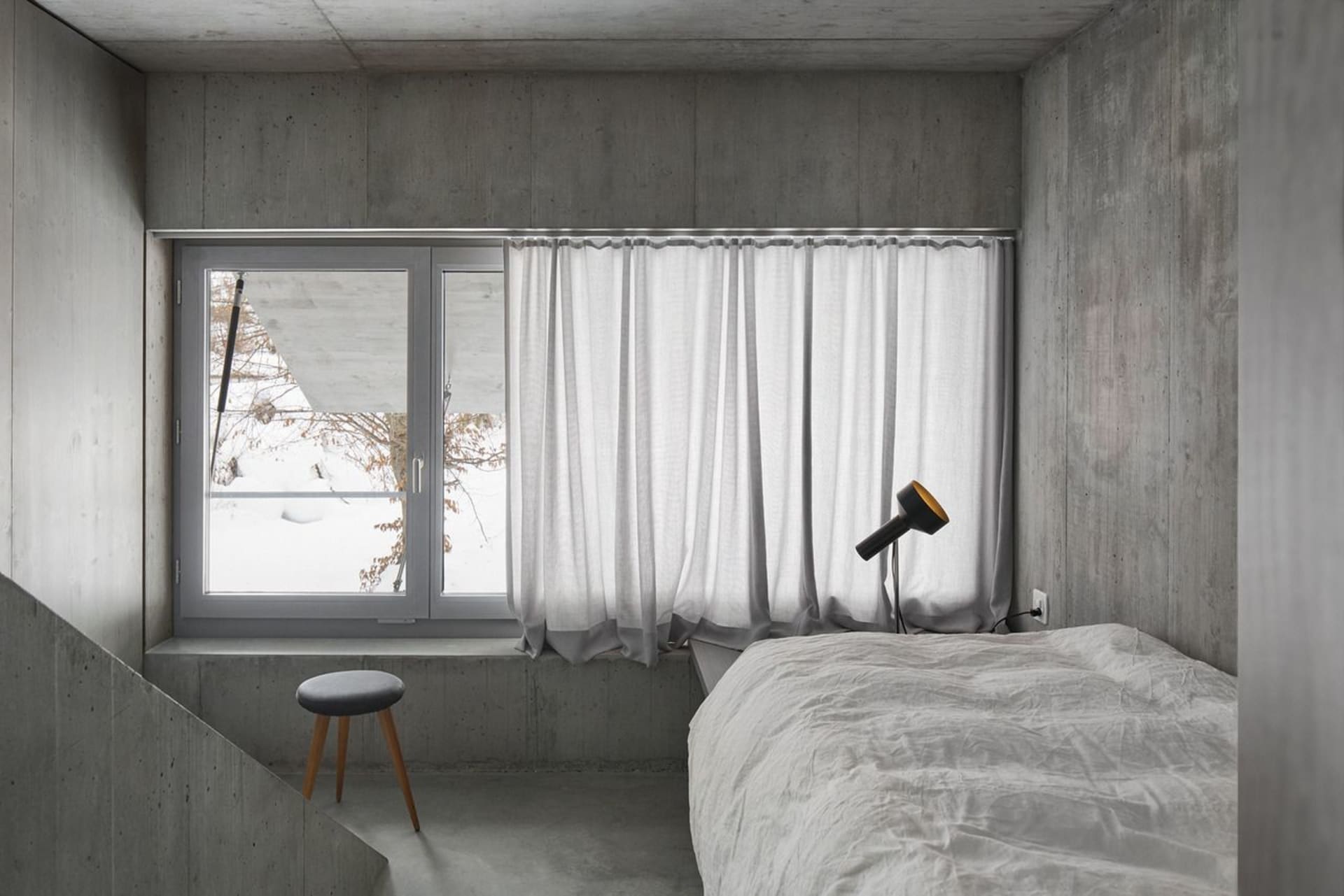 A minimalist bedroom with concrete walls, a white bed, and a white curtain at the window.