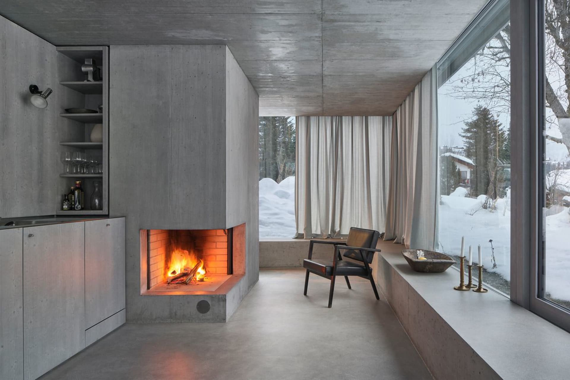 A modern living room with a concrete fireplace, minimalist decor, and large windows offering a snowy landscape view.