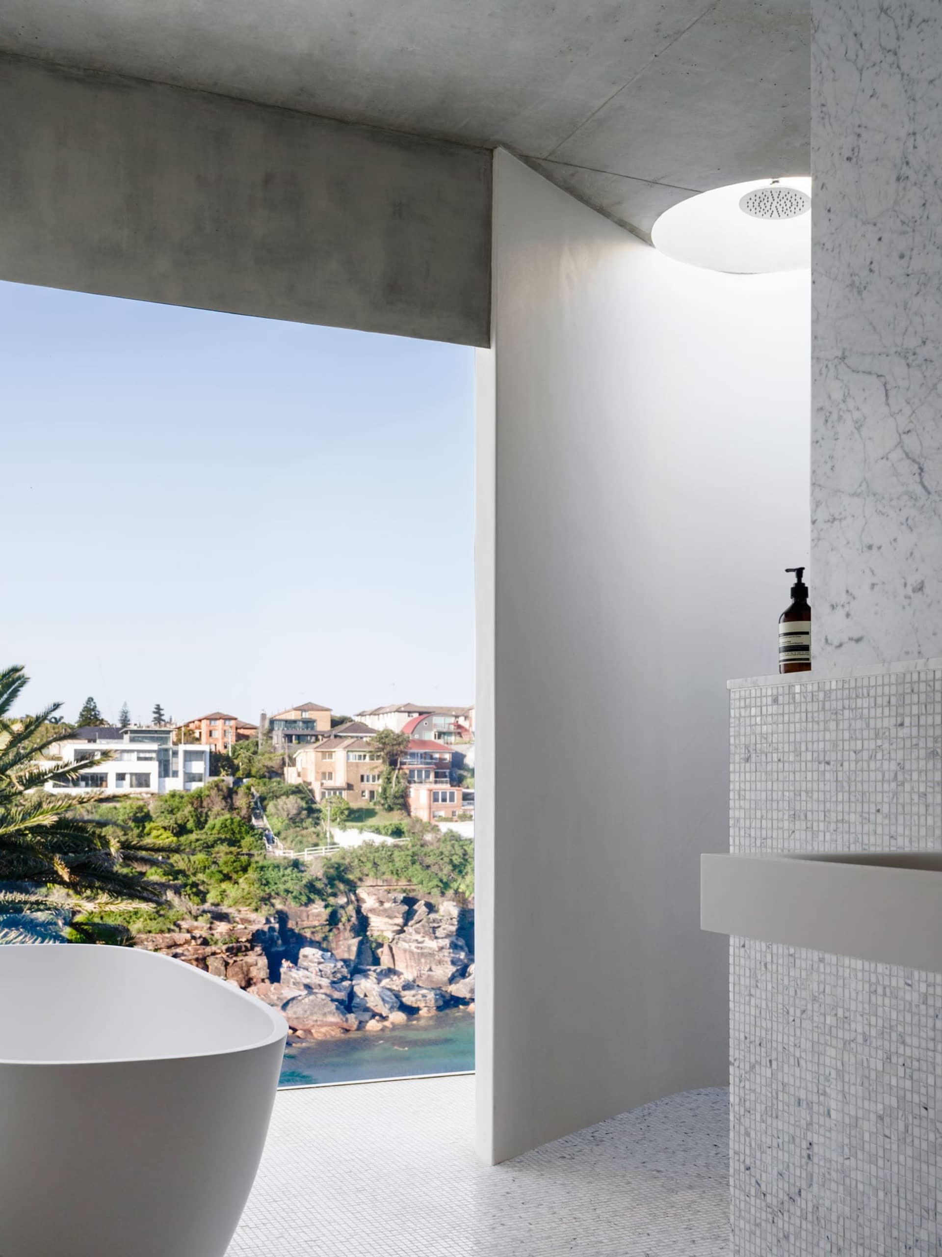 A modern bathroom with a freestanding concrete bathtub, marble tiles, and glass walls overlooking a coastal town.