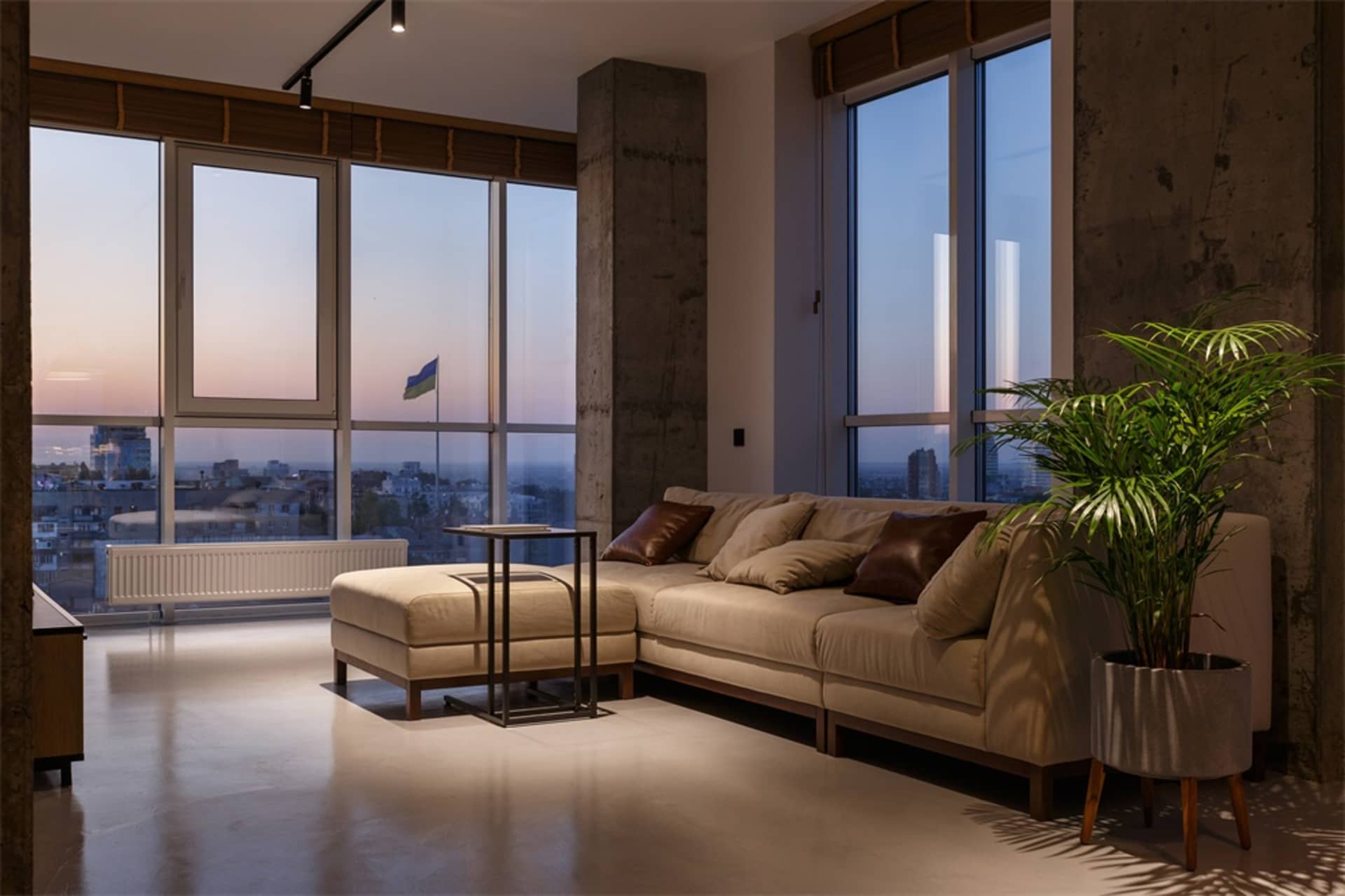 A modern living room featuring minimalist furniture and concrete flooring with a view of the city at dusk.