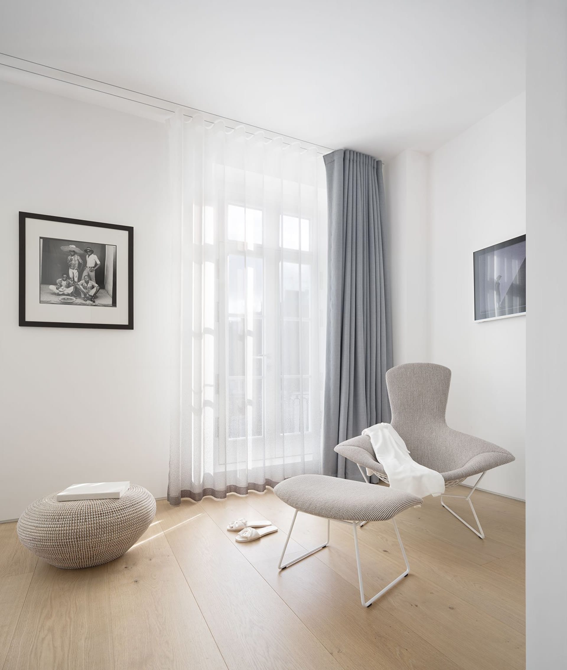 A minimalist living room featuring a grey upholstered chair, a white ottoman, and a glass window with sheer curtains.