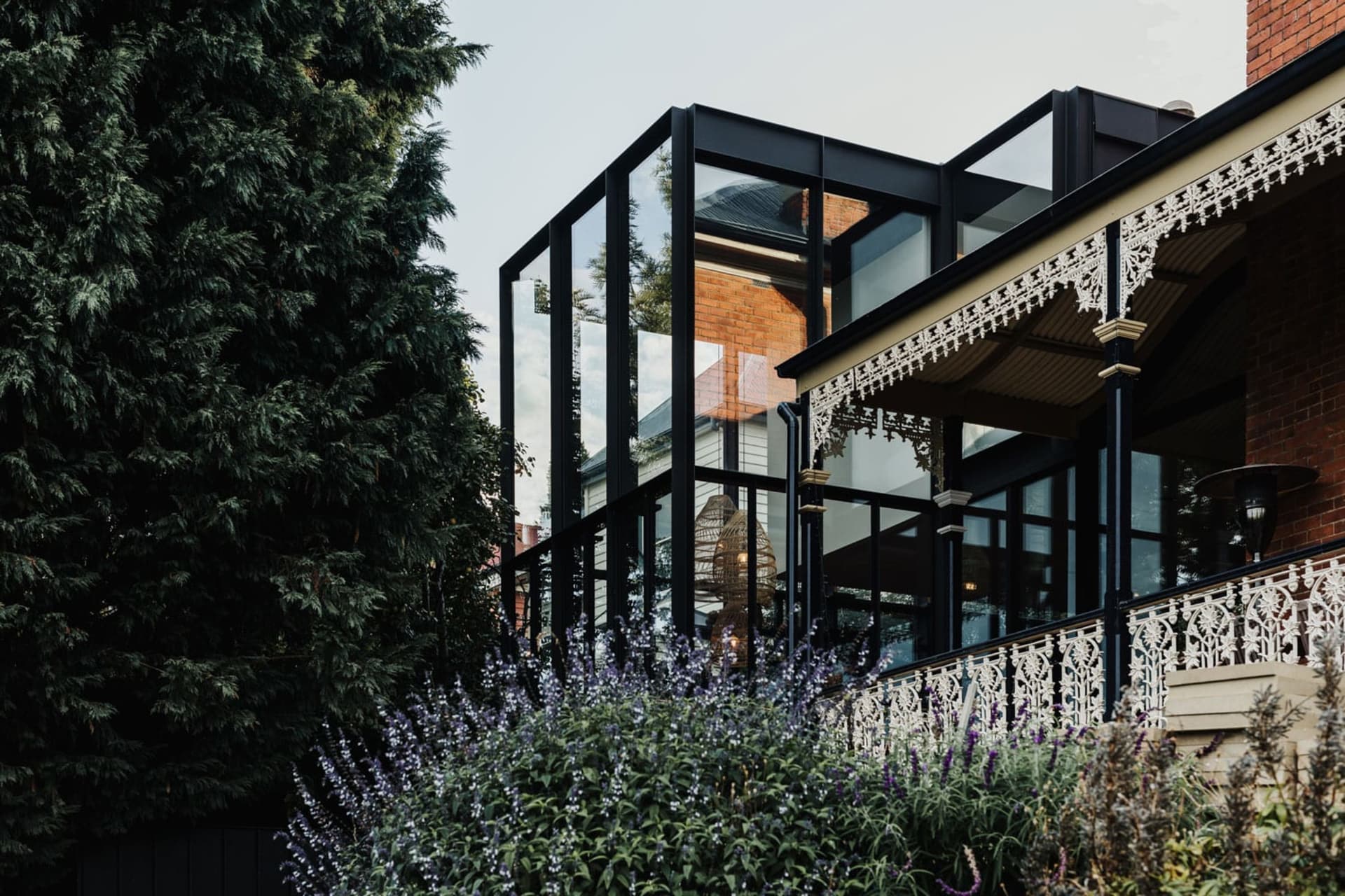 A modern house with large glass windows and a brick facade surrounded by lush greenery and purple flowers.