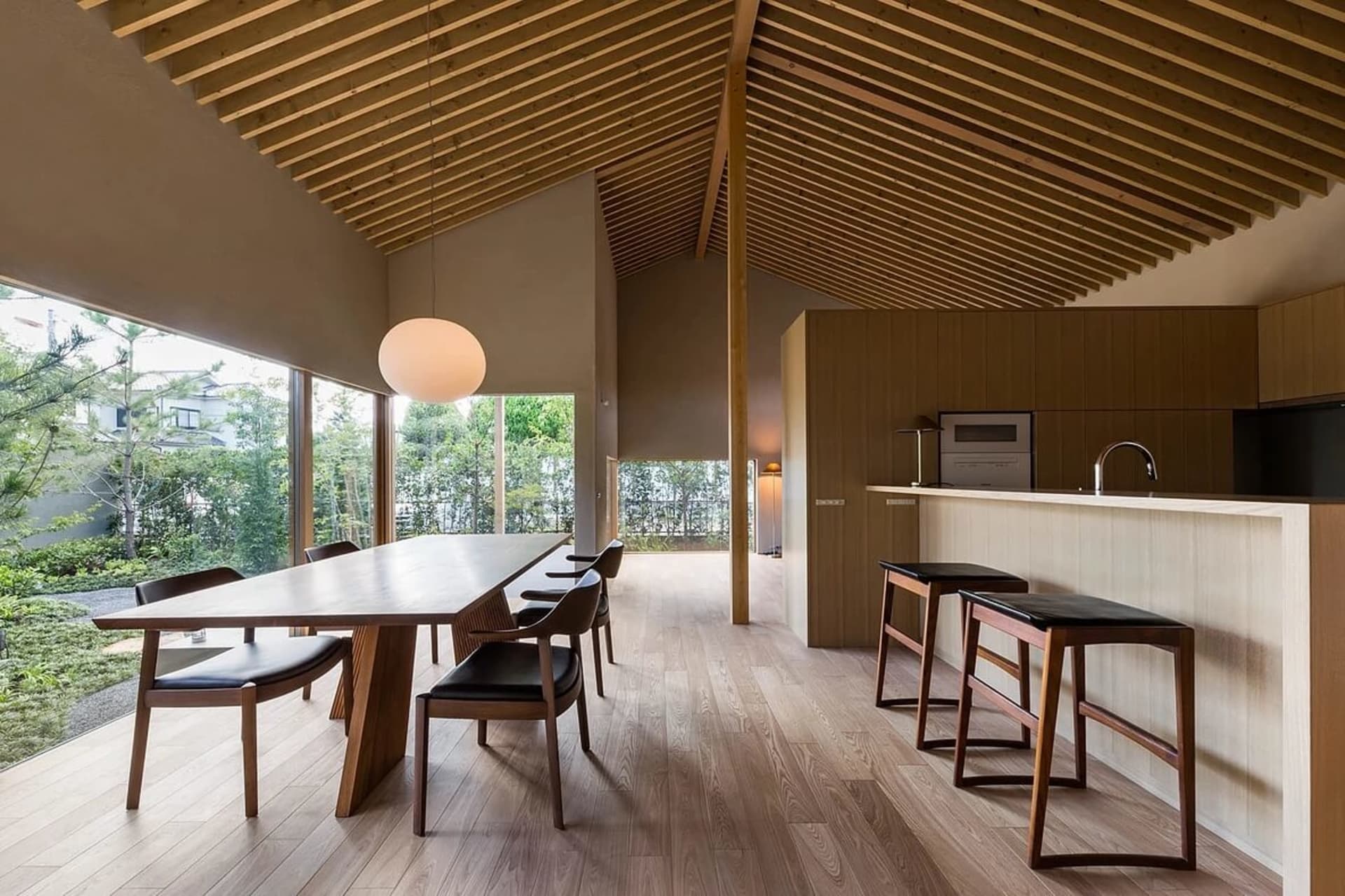 Modern minimalist kitchen with wood flooring and countertops, featuring black chairs and a long table.