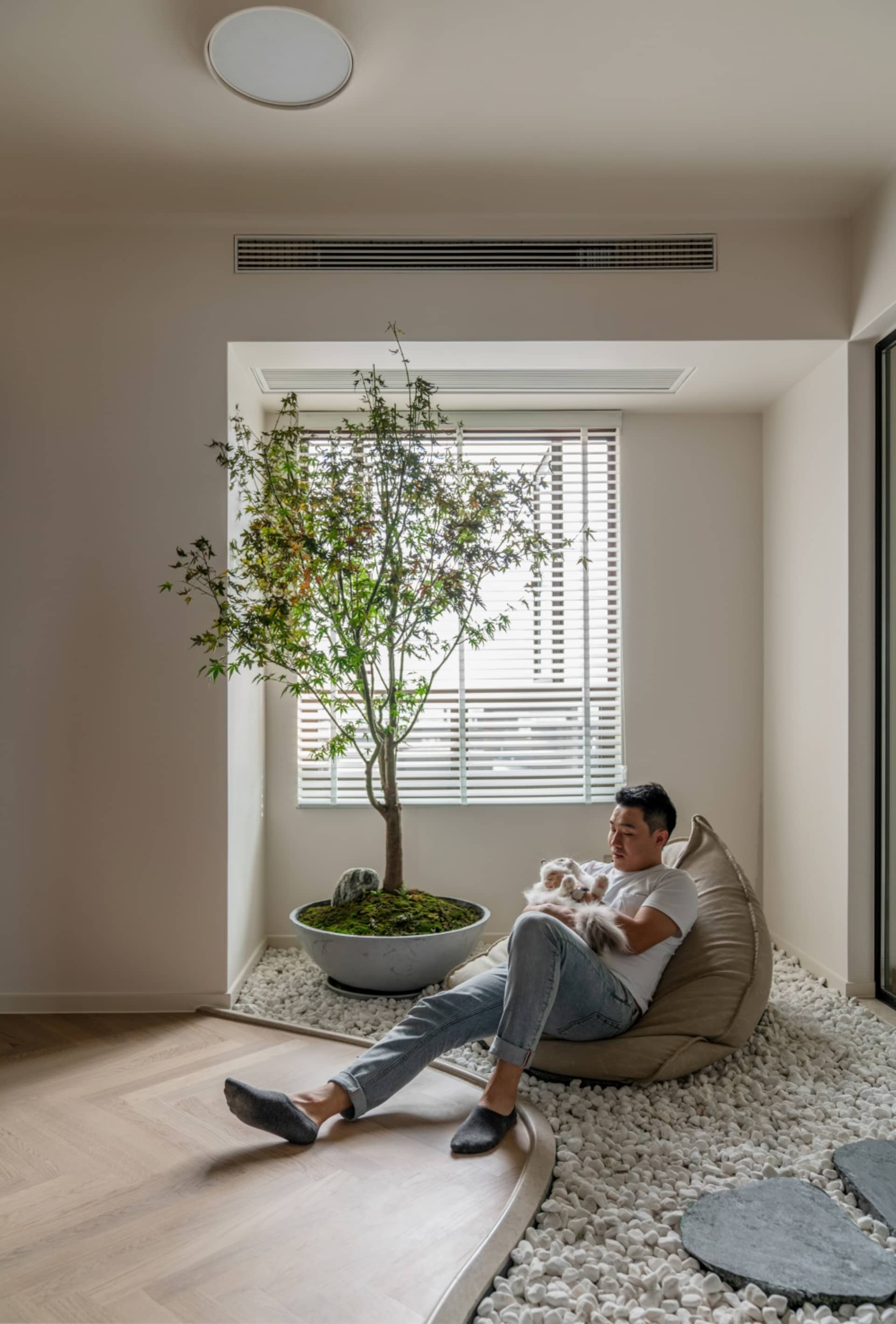 A serene modern living room featuring a bonsai tree in a moss-filled pot, pebbles on the floor, and a man sitting on a cushio