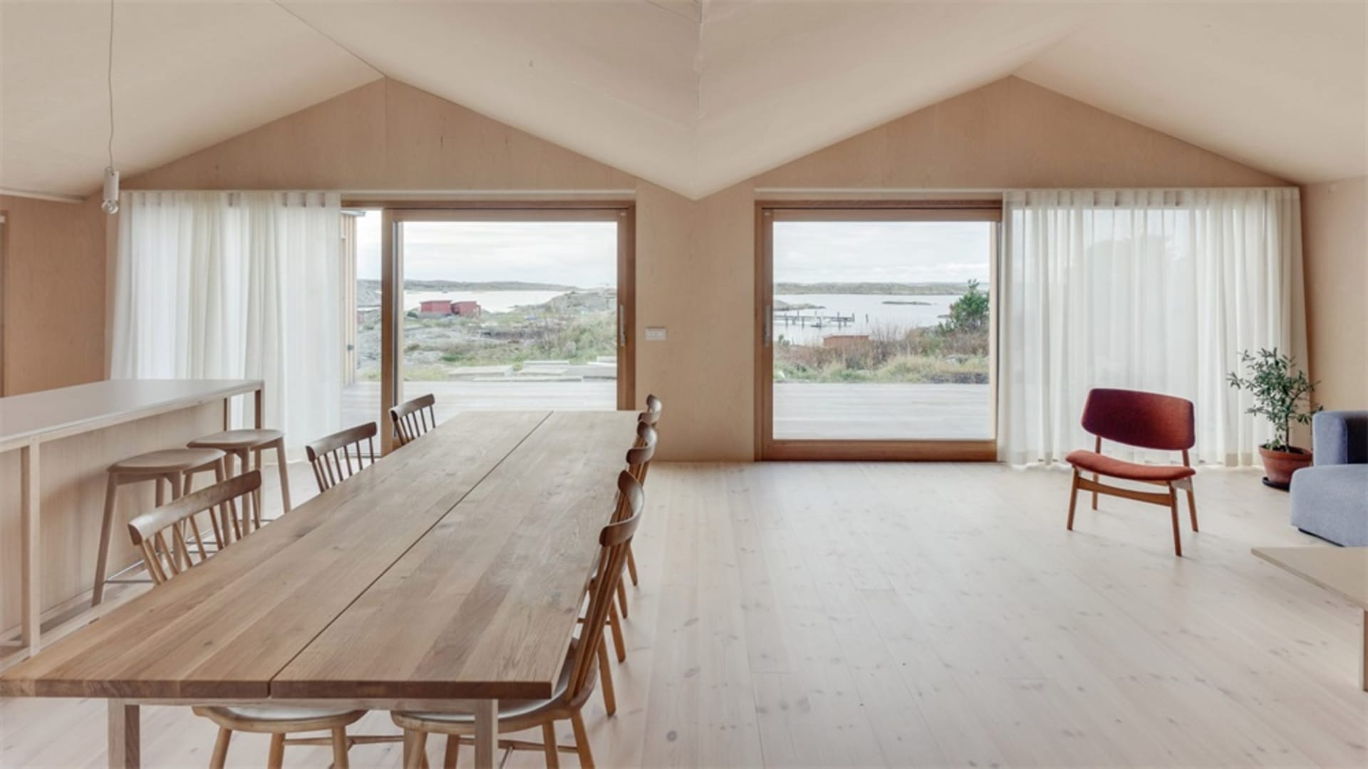 A modern residential living room with a high ceiling, large windows, and a long wooden dining table.