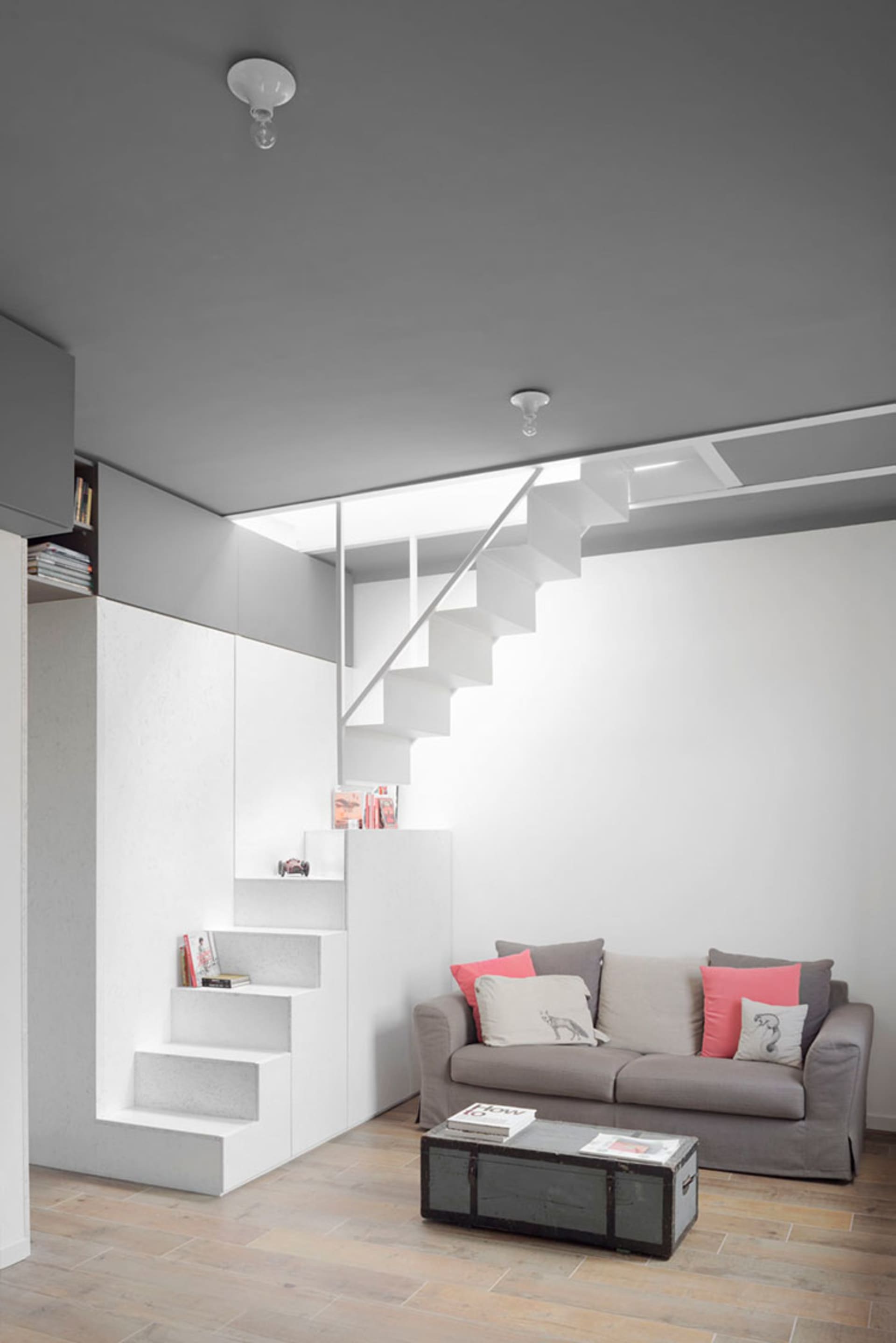 A modern minimalist living room with stairs, concrete flooring, and a gray sofa with pink and white cushions.