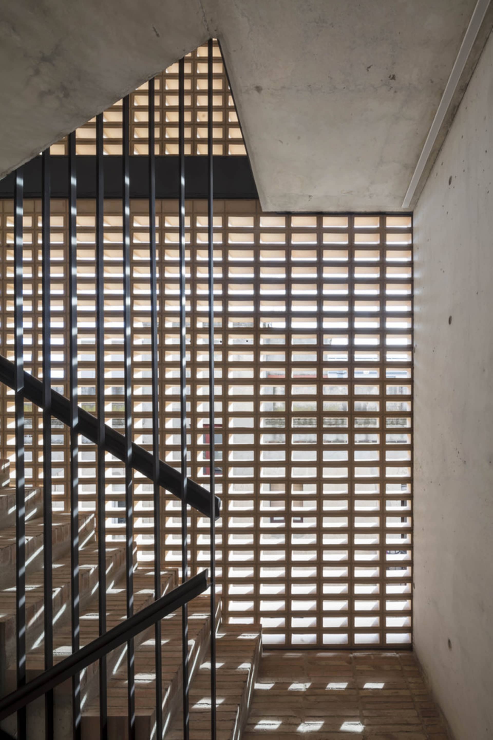 A modern staircase featuring a grid of wooden slats with concrete walls and black metal railings, illuminated by sunlight.