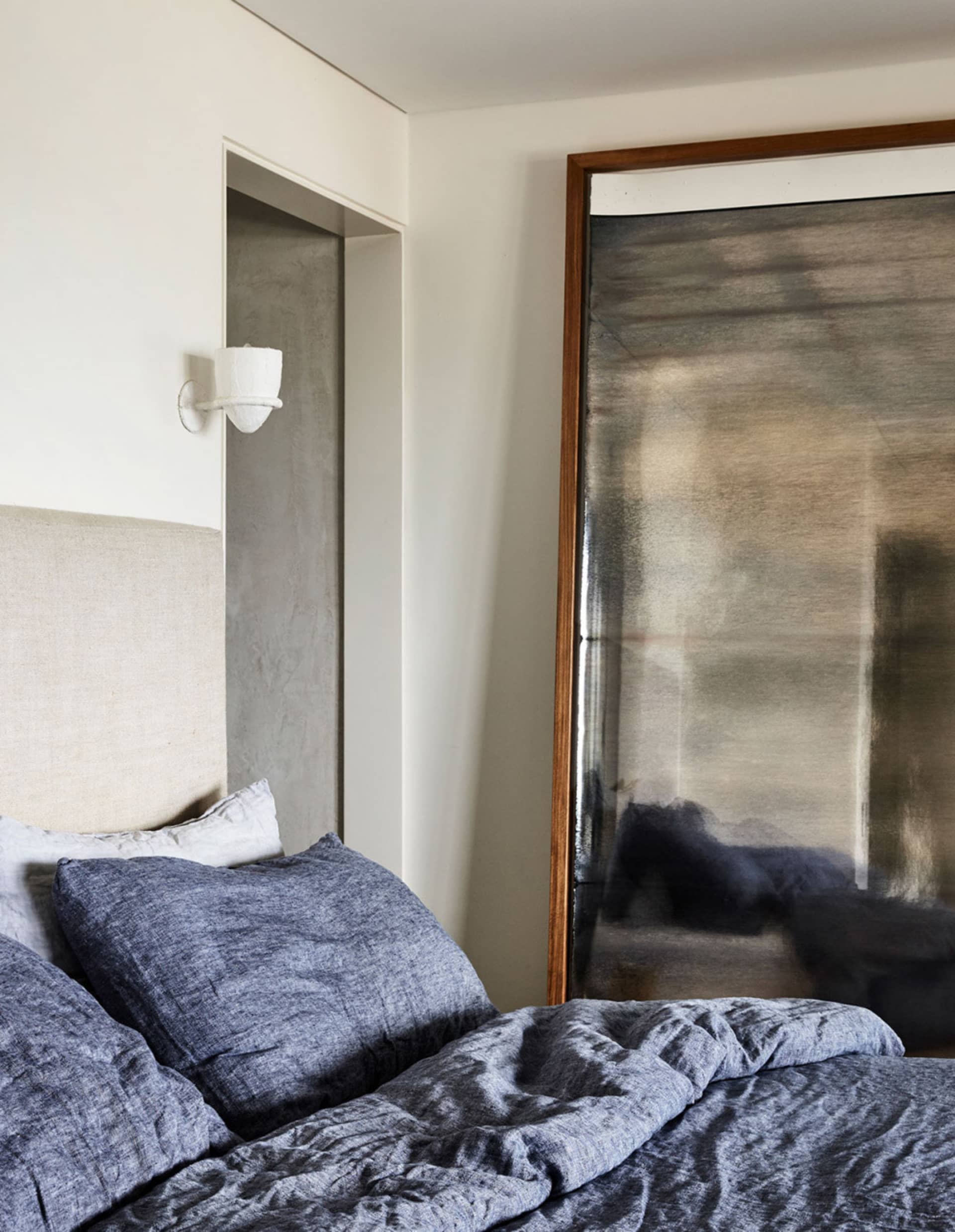 A minimalist bedroom with textured blue bedding and a neutral headboard against a white wall, featuring a large mirror with a