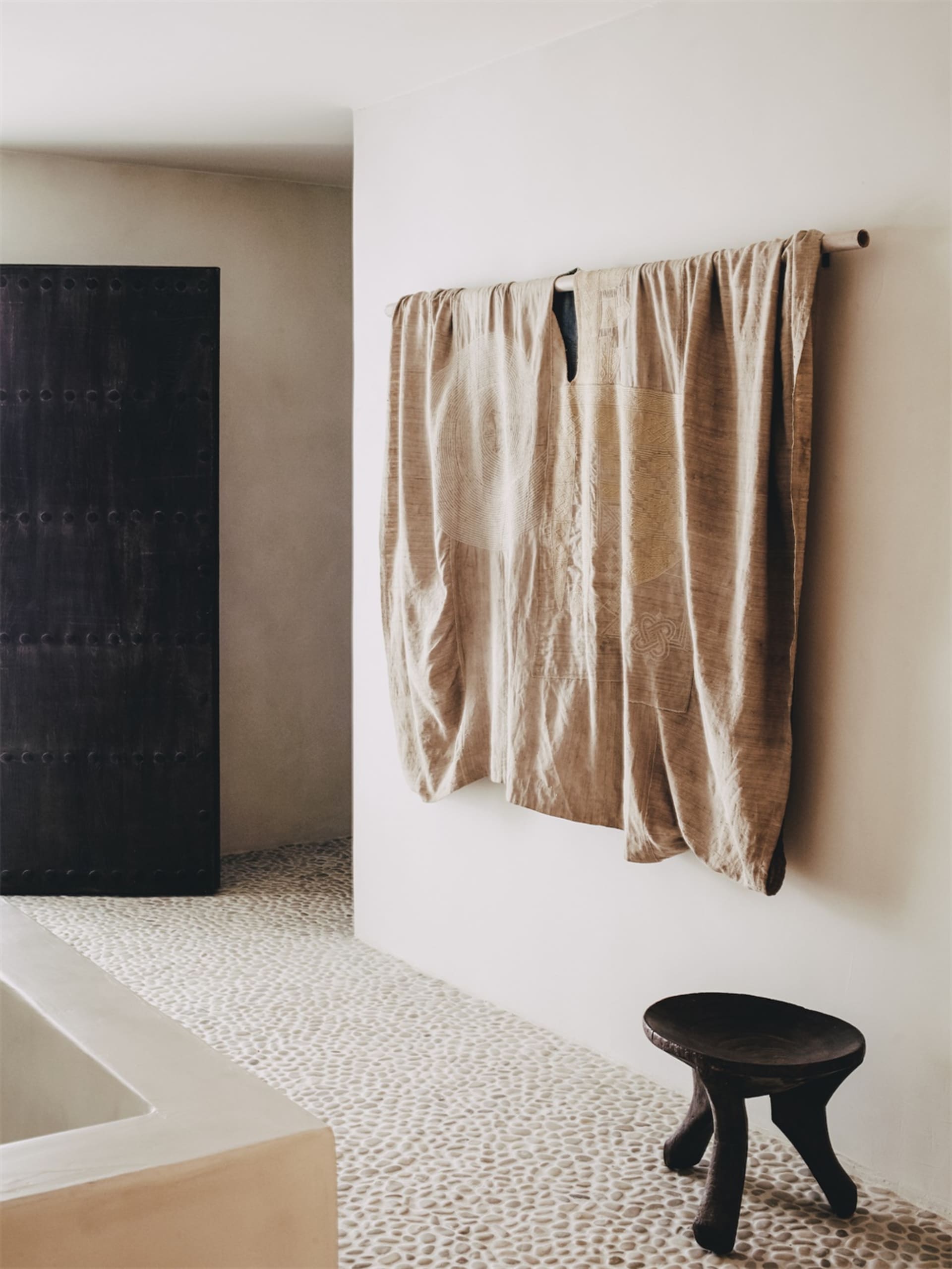 A textured, beige silk wall hanging in a modern bathroom with white pebble tile flooring and a large black framed mirror.