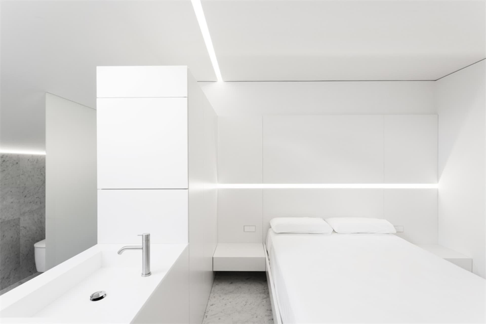 A modern minimalist bedroom with white furniture, a bed with white bedding, and a white sink with a mirror.