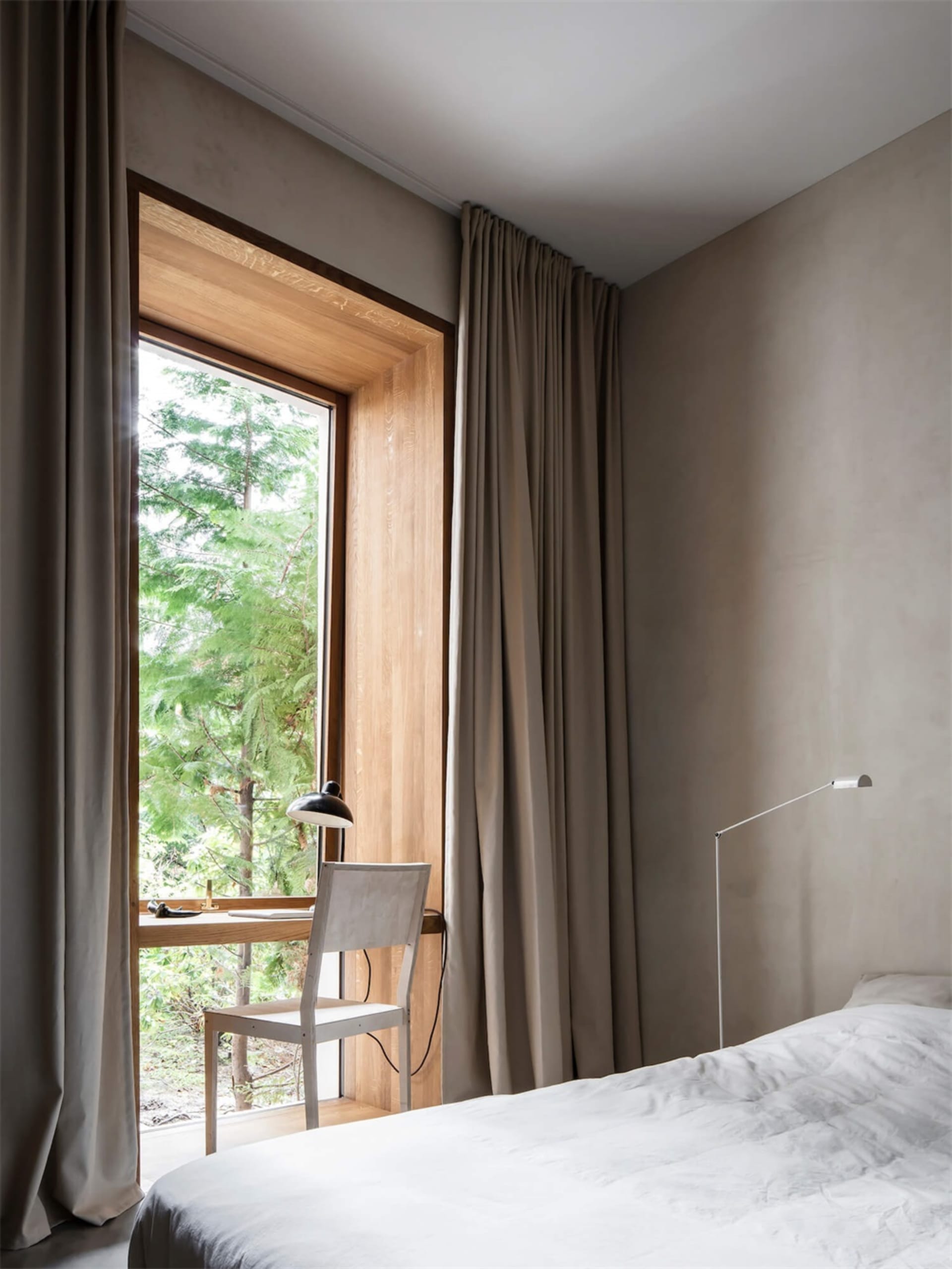 A minimalist bedroom with neutral-colored walls, curtains, and bedding. A small desk with a lamp and chair is placed next to 
